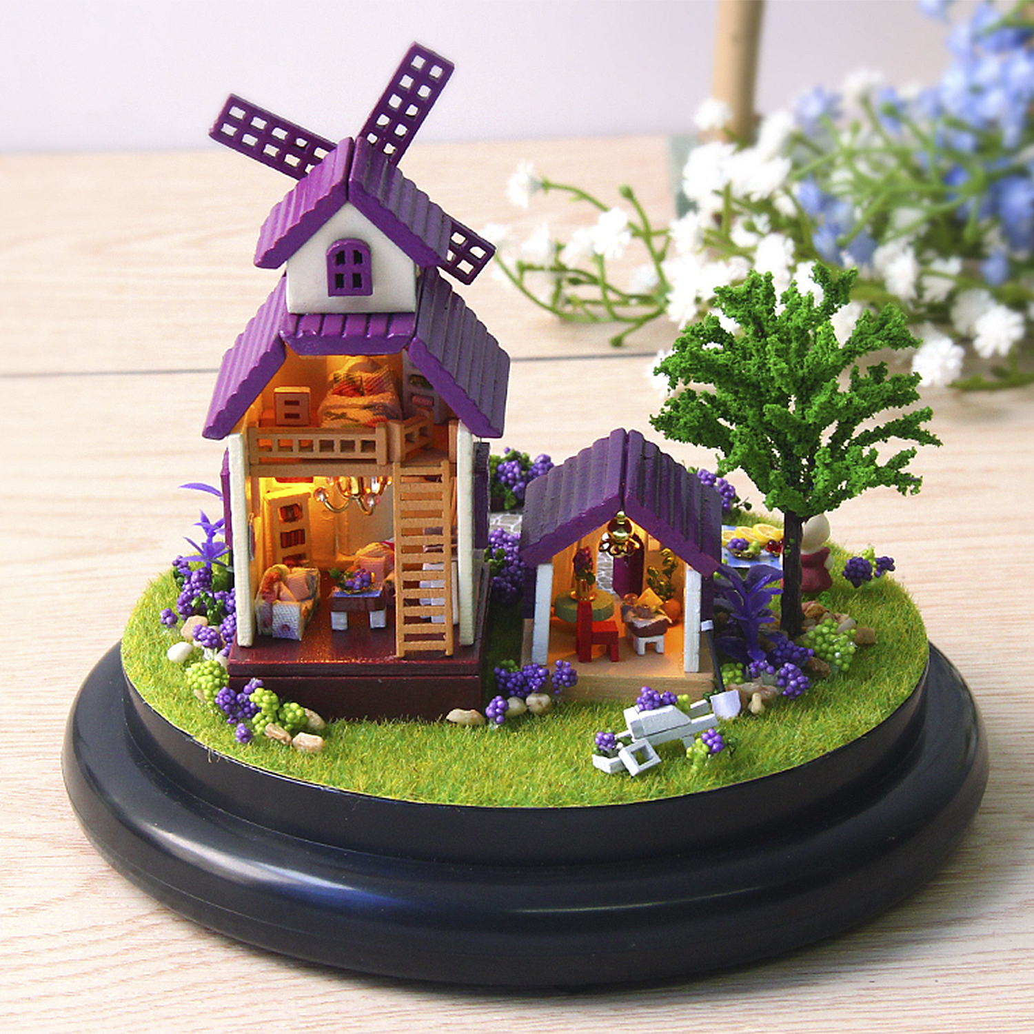 Mini mulino a vento con casa, albero e fiori su base erbosa.