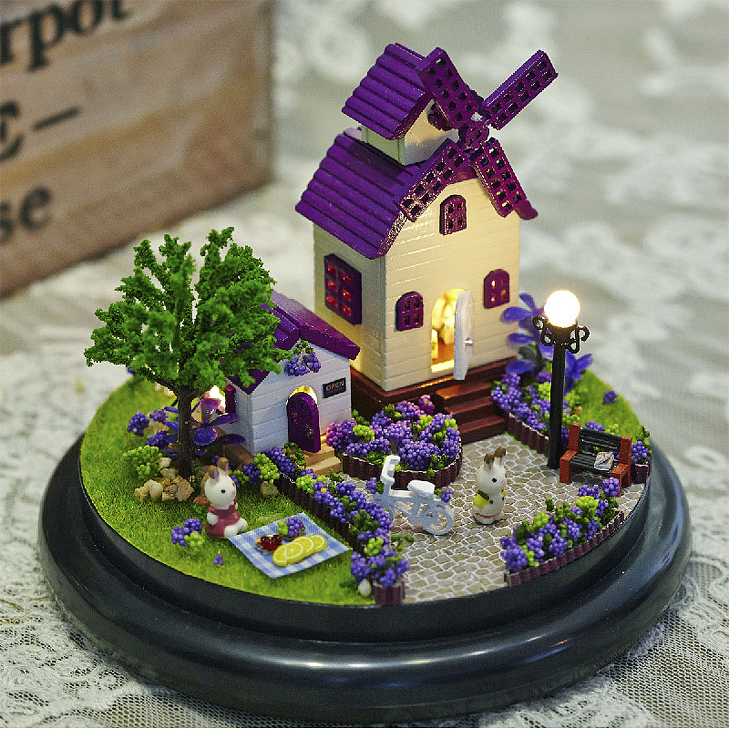 Villaggio in miniatura con mulino, casa, albero, conigli, bici, panchina.
