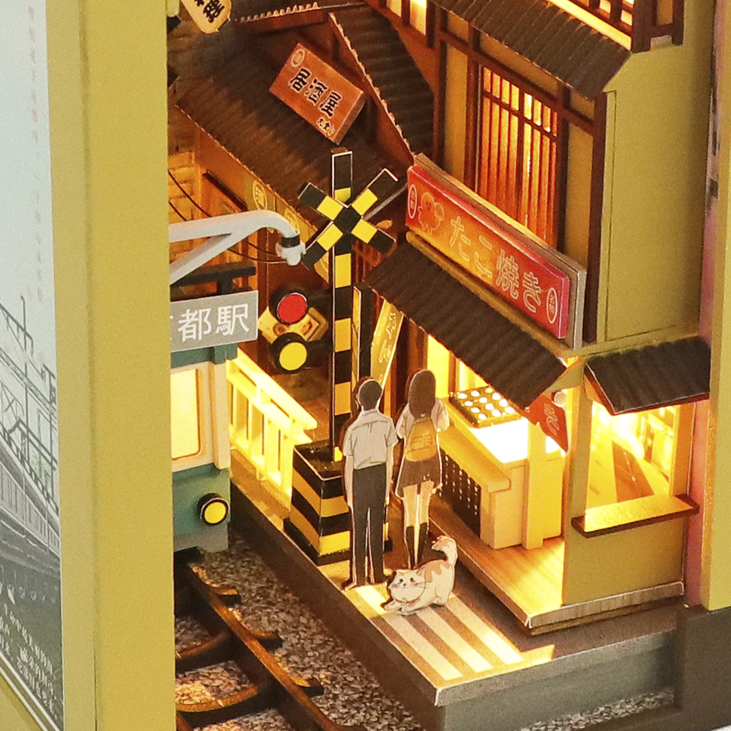 Scena in miniatura di stazione ferroviaria con treno, figure, gatto e negozi illuminati.