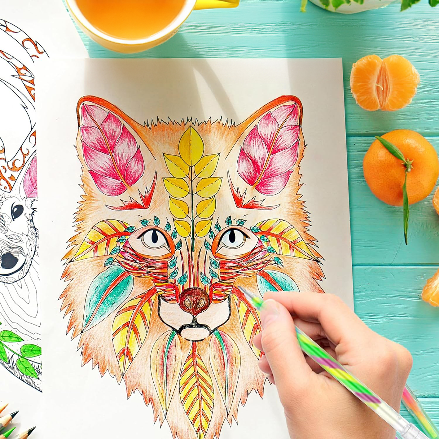 Una mano colorea un zorro con bolígrafos de colores sobre papel junto a té y mandarinas.