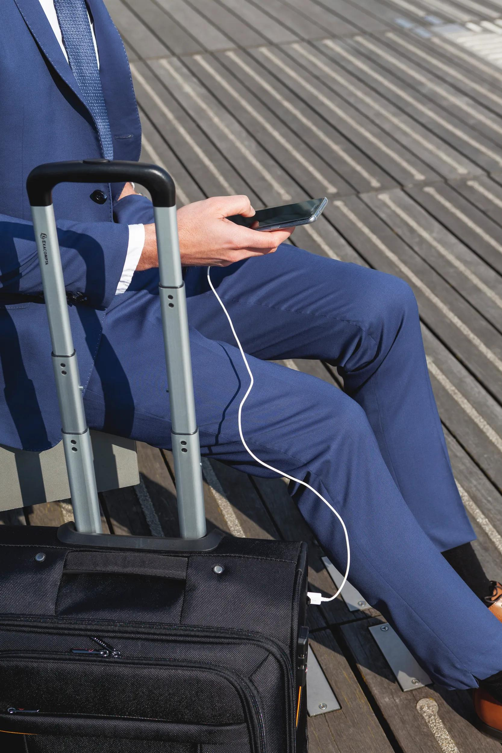 Hombre con pantalón y chaqueta azul usa smartphone cargando desde maleta.