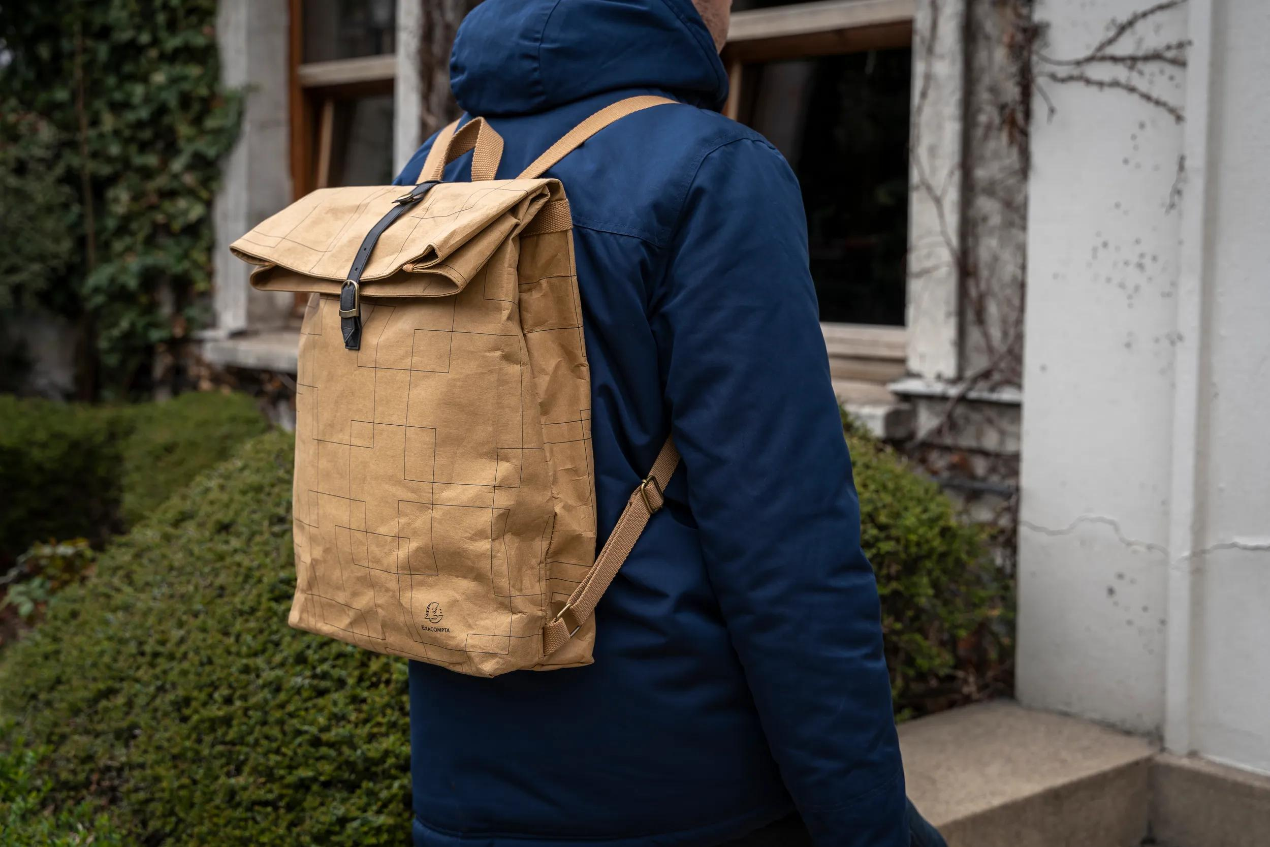 Persona con mochila marrón y chaqueta azul frente a una casa con plantas.