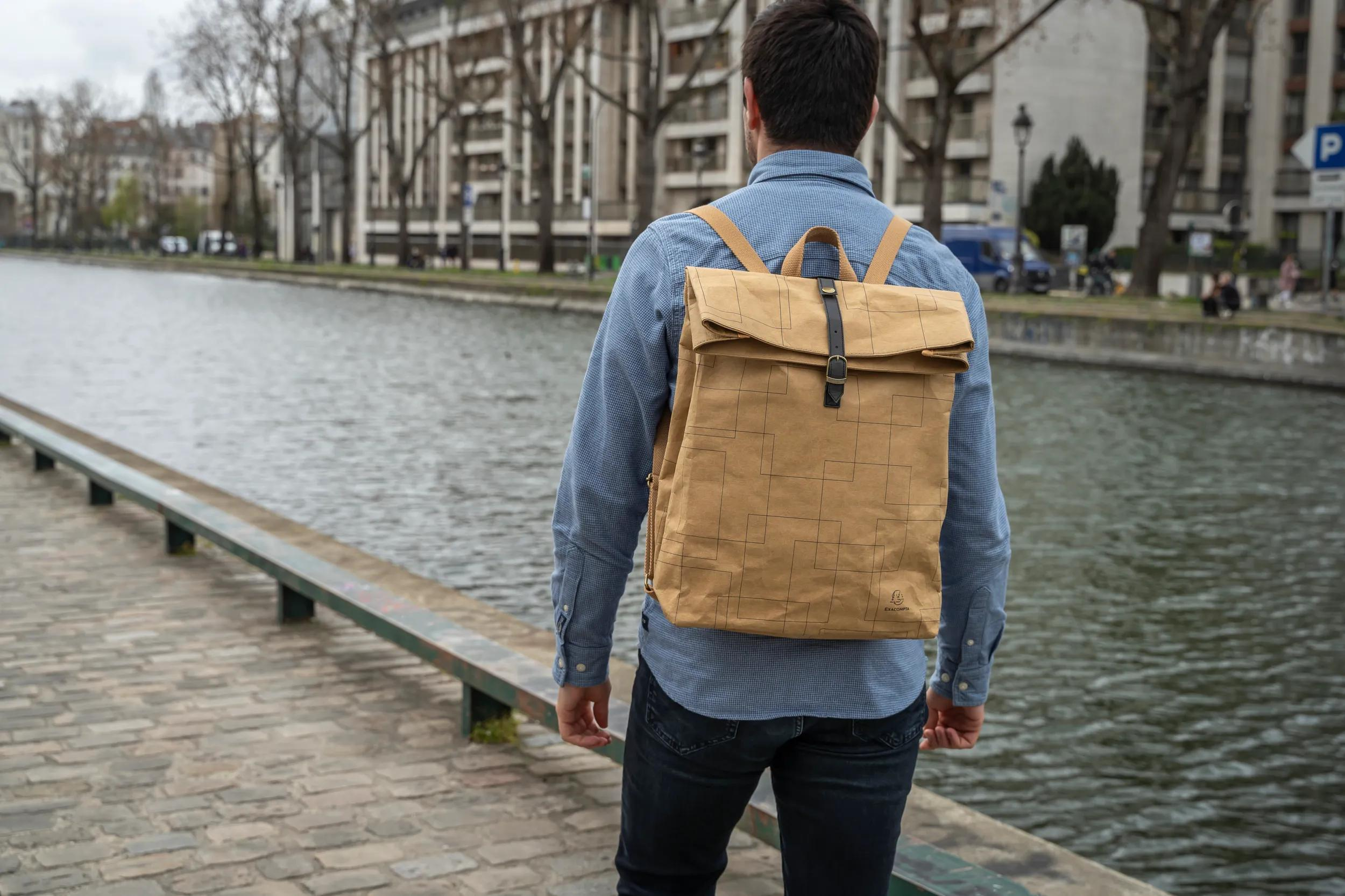 Hombre con mochila marrón junto al canal, edificios al fondo.