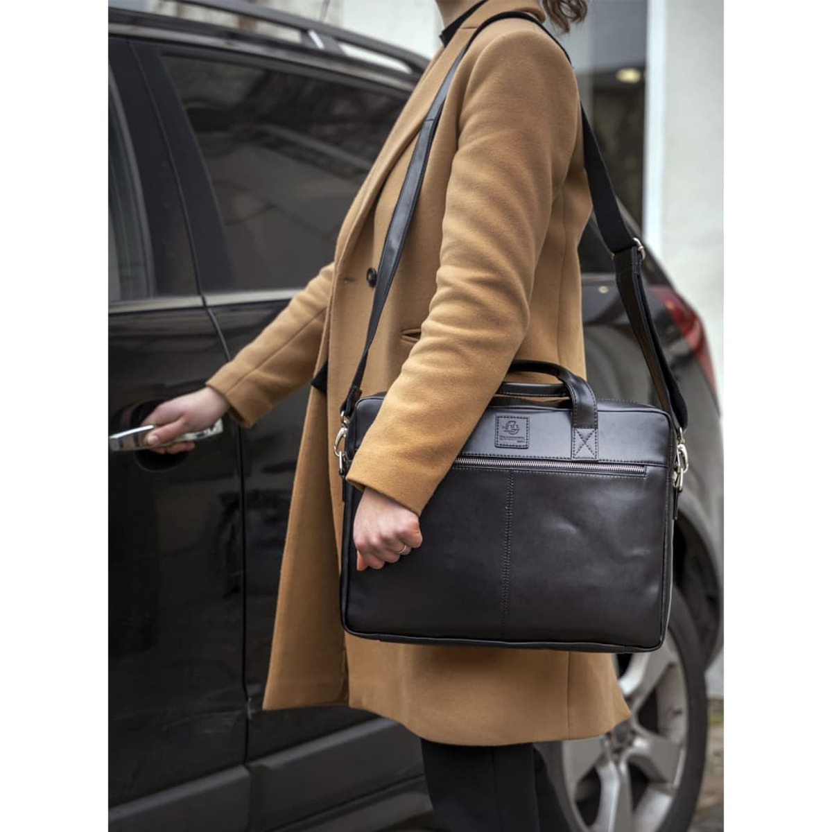 Una persona con abrigo color camello abre la puerta de un coche, llevando un bolso de cuero negro.