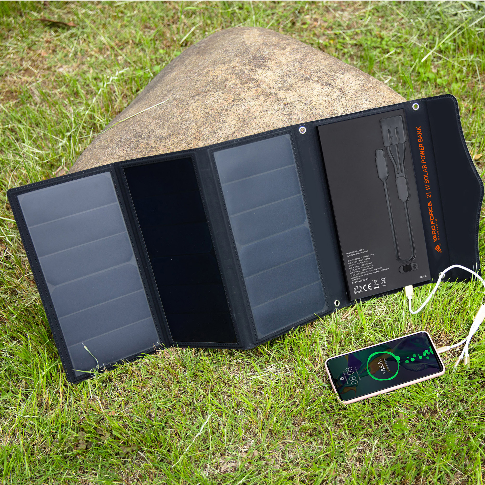 Ein faltbares Solar-Ladegerät mit Kabeln und einem Handy, das auf Gras liegt.