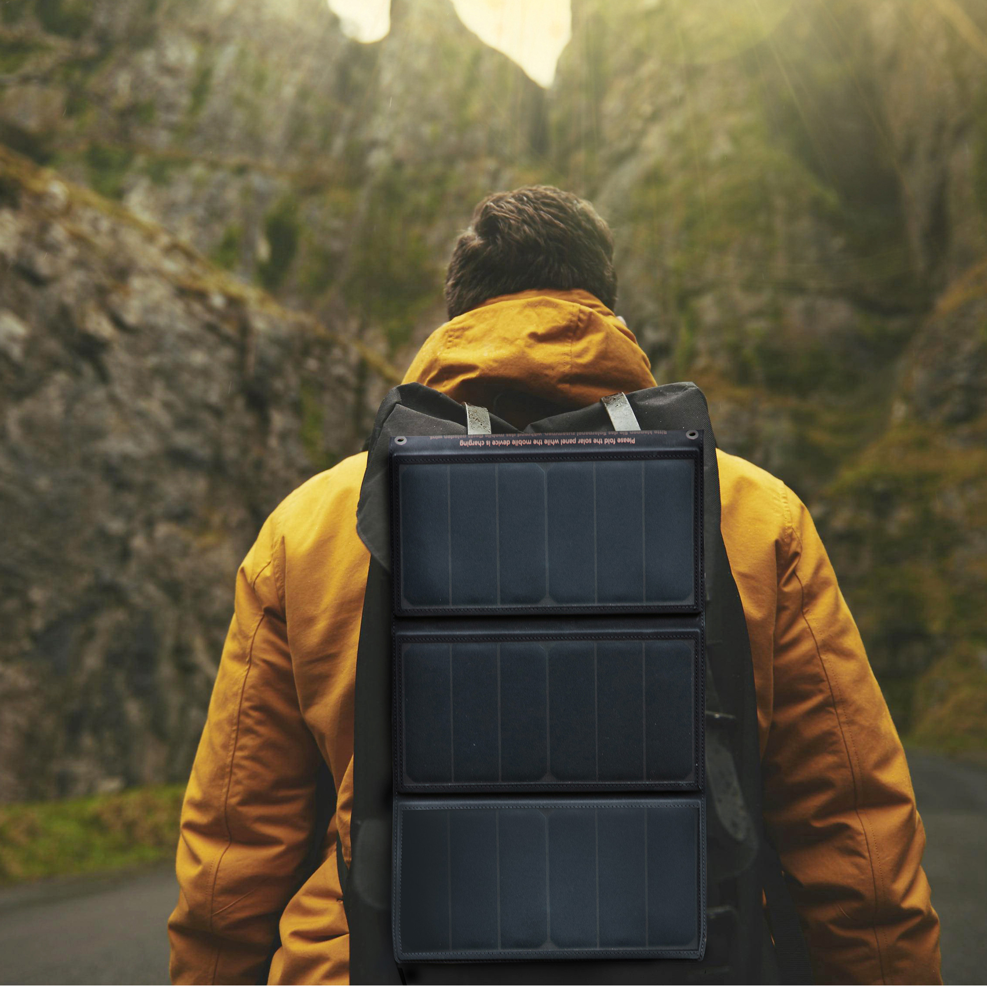 Mann in gelber Jacke mit Solarpanel-Rucksack in Berglandschaft.