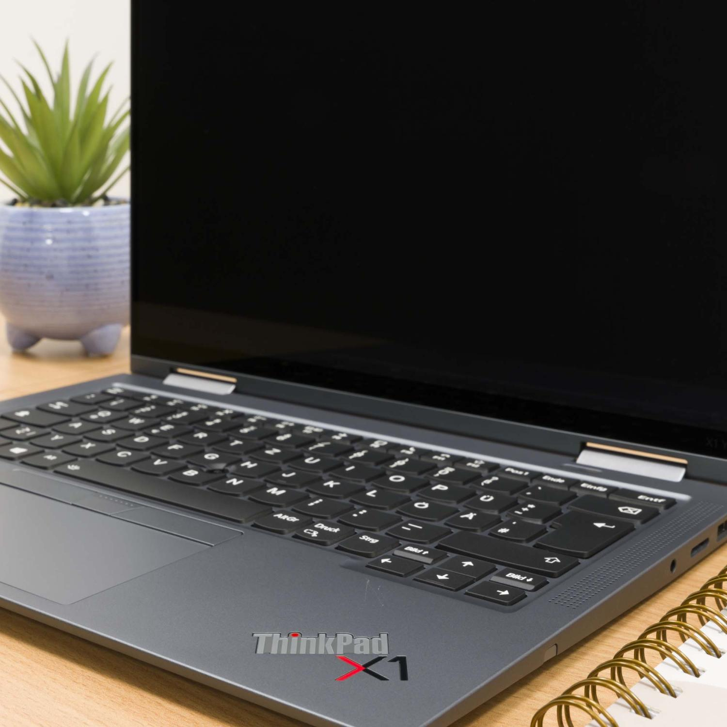 Ein geschlossener Laptop mit ThinkPad X1 Logo, neben einer Topfpflanze und Notizbuch.