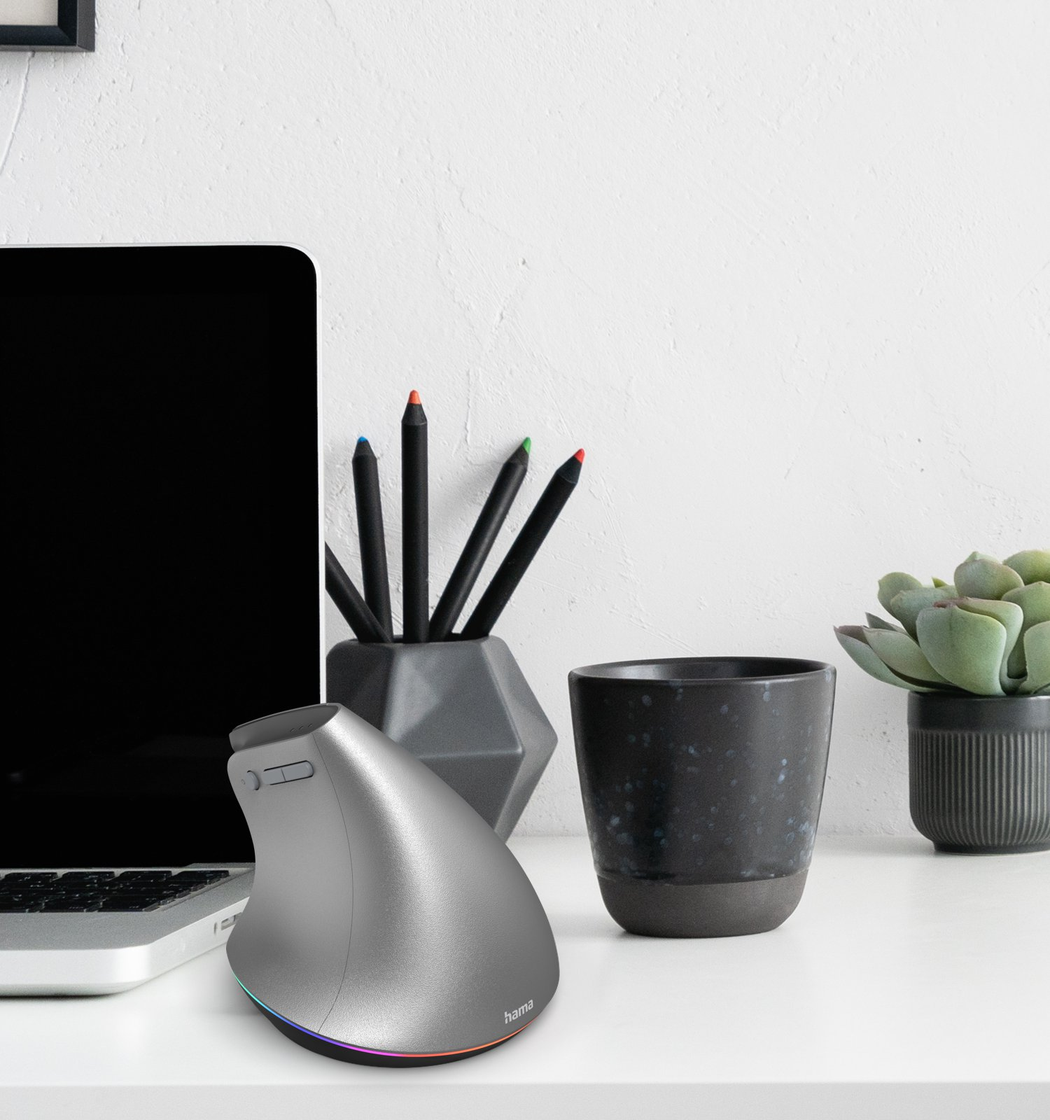 Szara pionowa mysz obok laptopa, pisaków, kubka i rośliny.