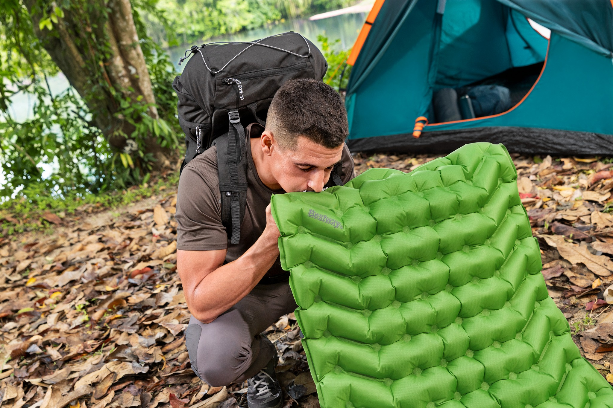 Ein Mann mit Rucksack bläst eine grüne Isomatte auf, Zelt im Hintergrund.