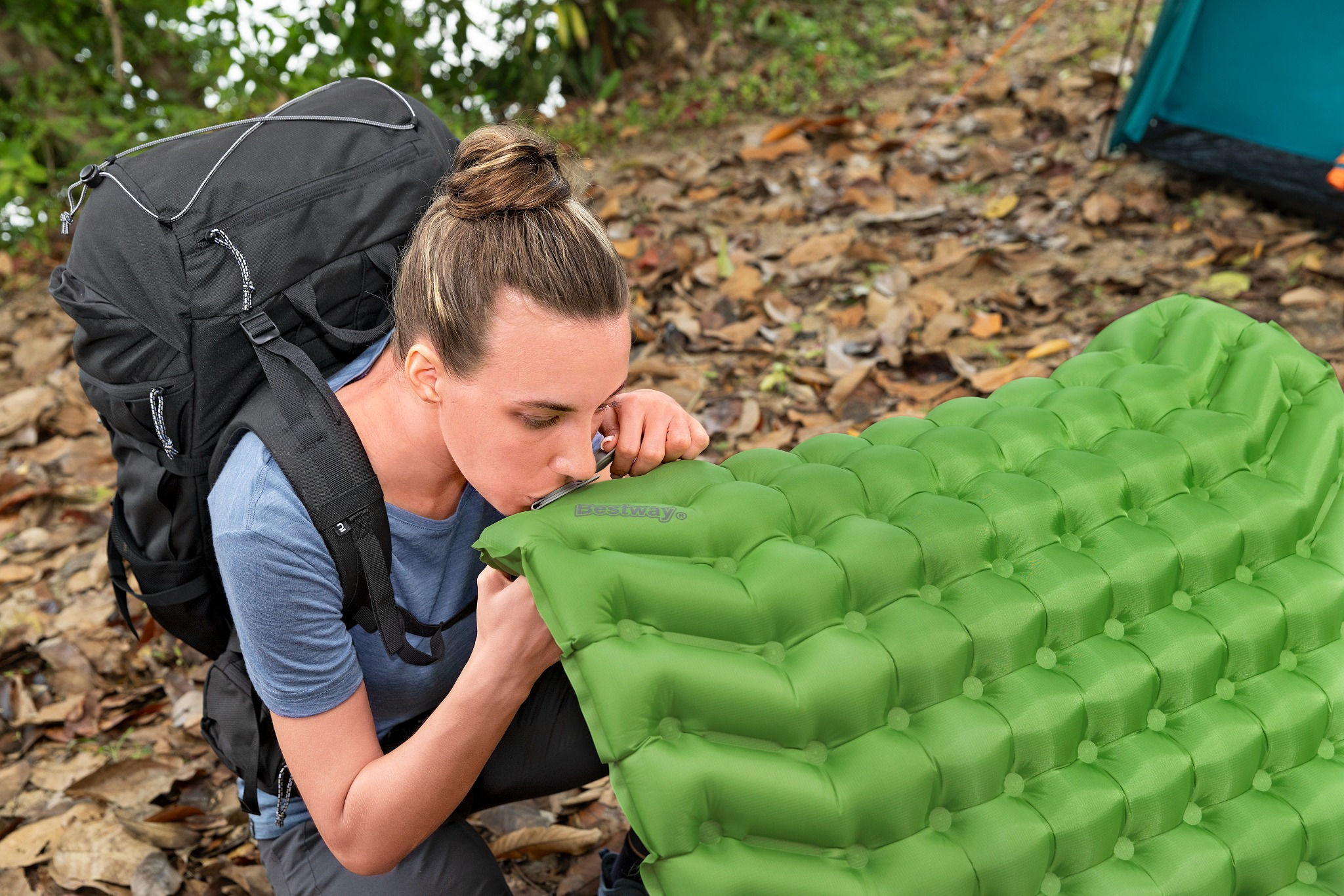 Eine Frau mit Rucksack bläst eine grüne Isomatte auf. Ein Zelt ist im Hintergrund.