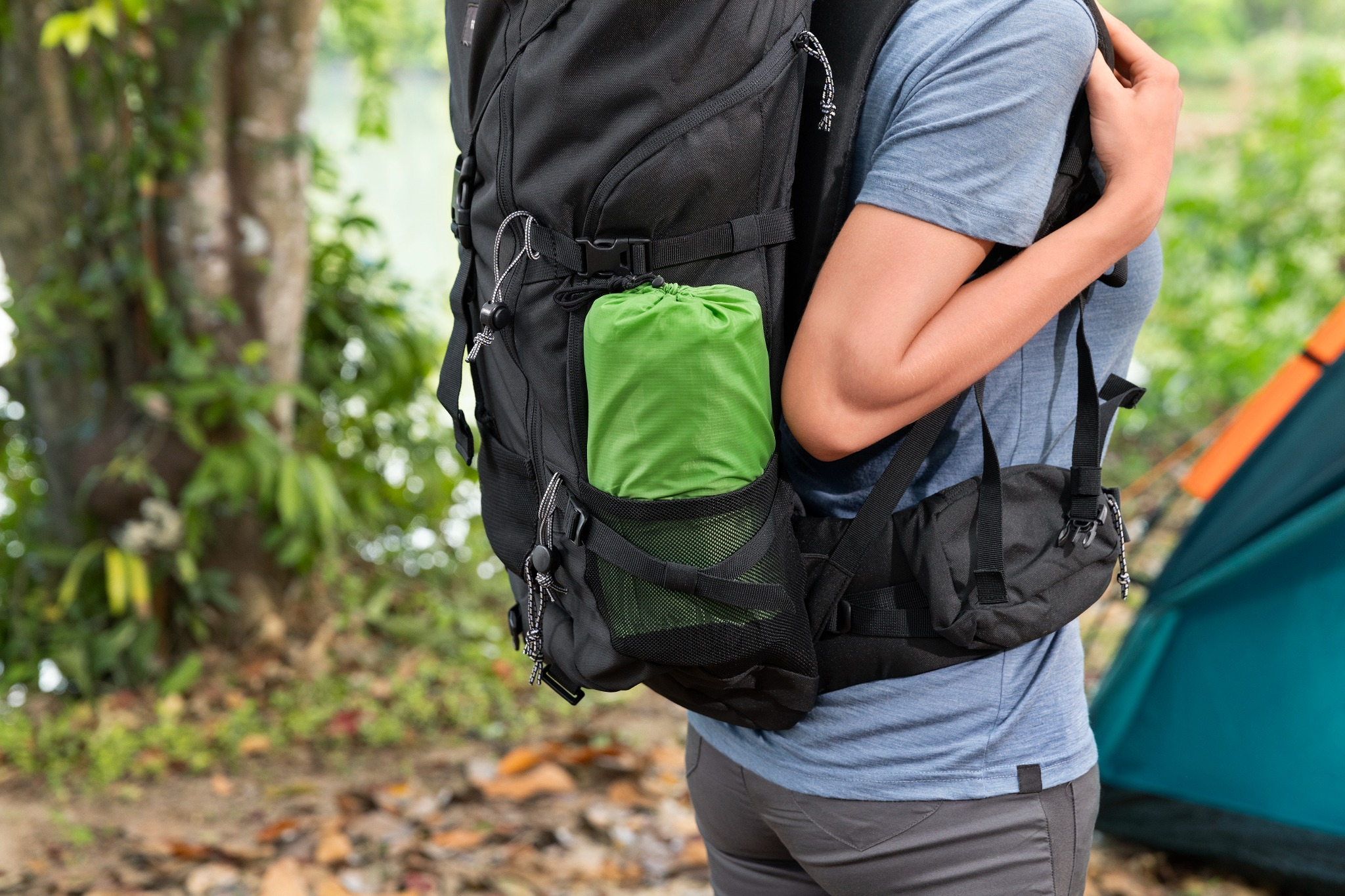 Wanderer mit Rucksack und grüner Rolle im Wald, Zelt im Hintergrund.