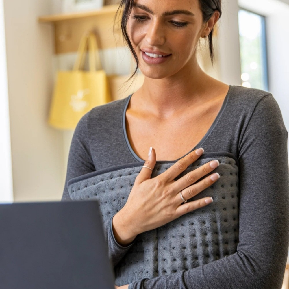 Woman holds heating pad, looks at laptop. Yellow bag in background.