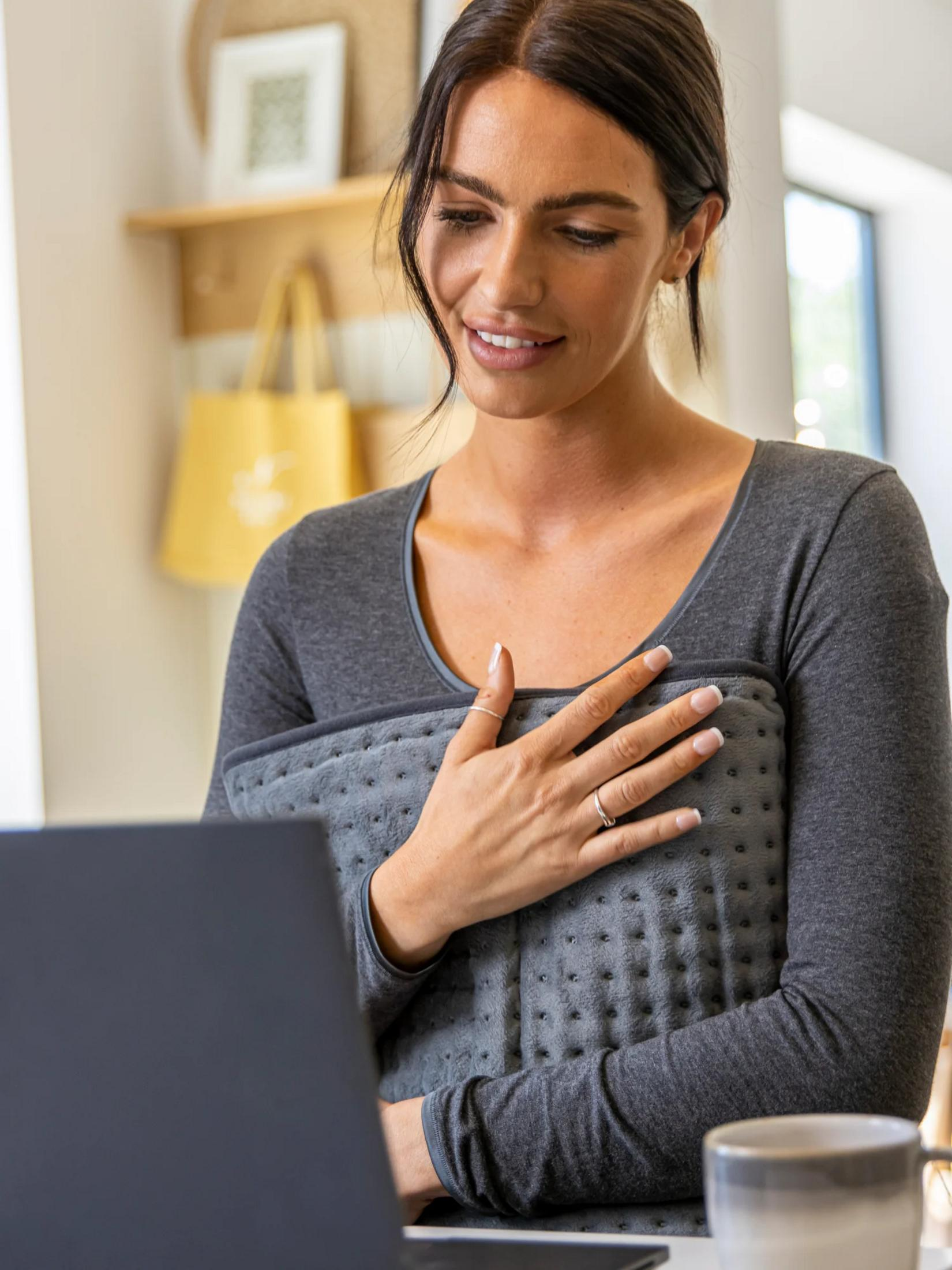 A woman holds a gray heating pad and looks at her laptop.