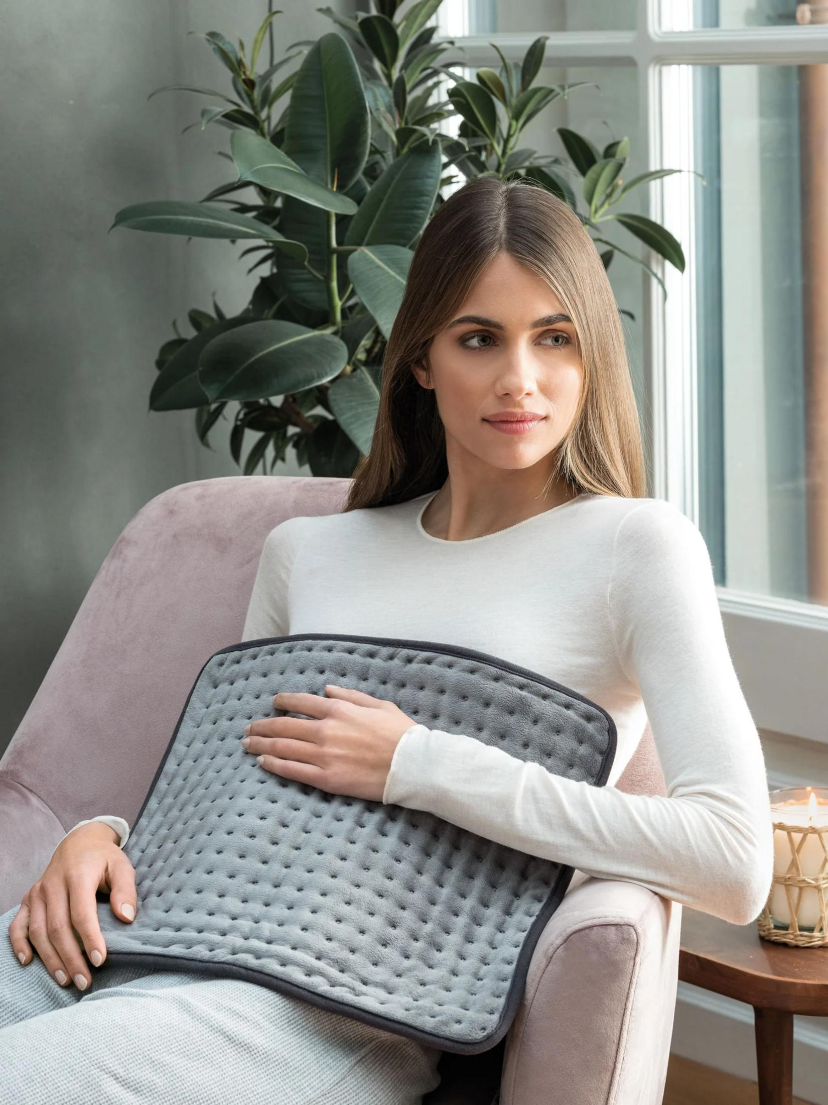 A woman sits in an armchair holding a grey heating pad on her lap.