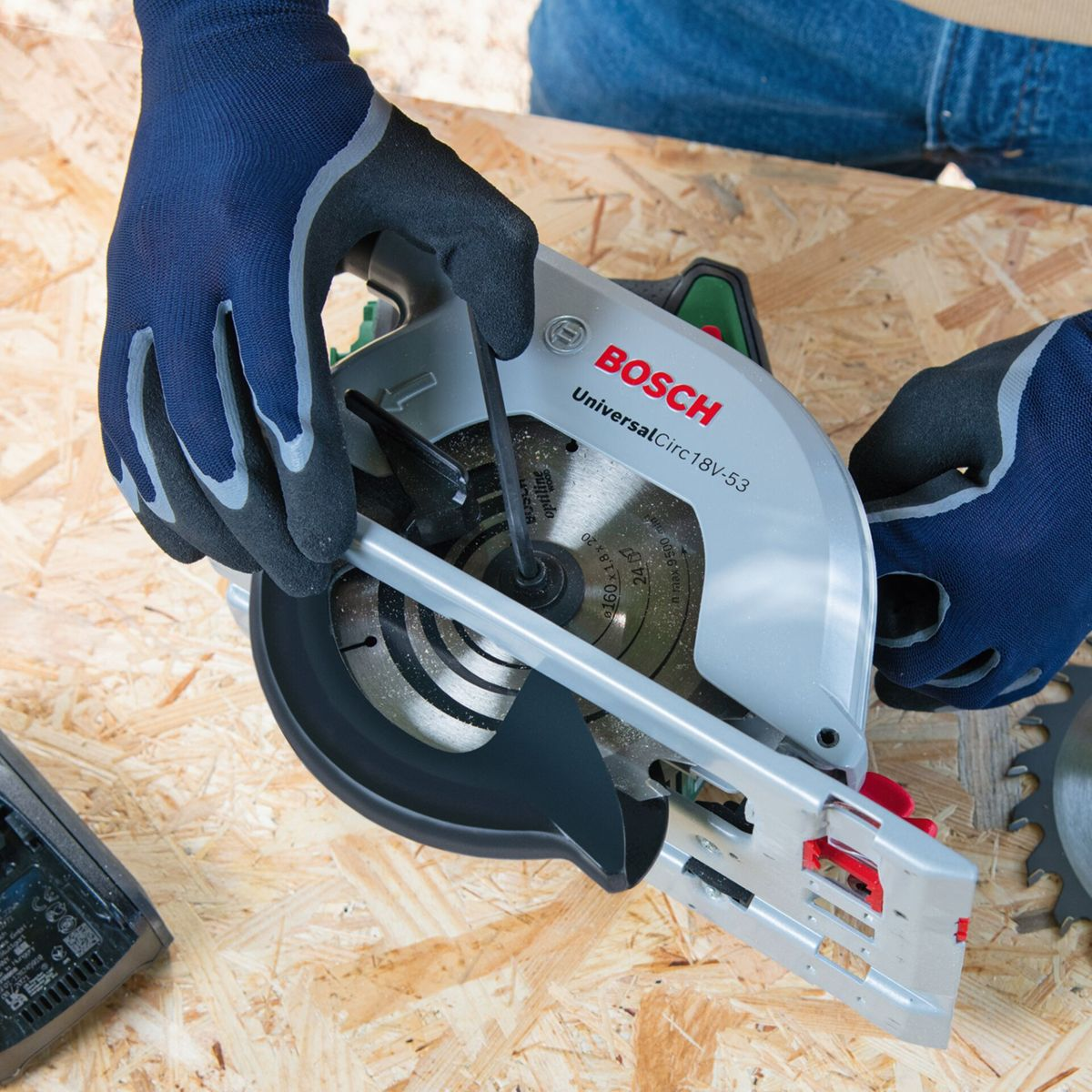 Hände in blauen Handschuhen halten eine Bosch Kreissäge über Holz.