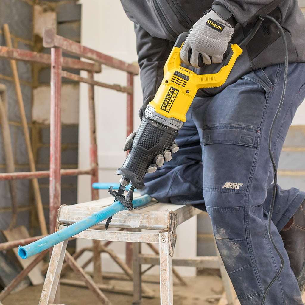 Man cuts pipe with yellow reciprocating saw. Construction work in background.