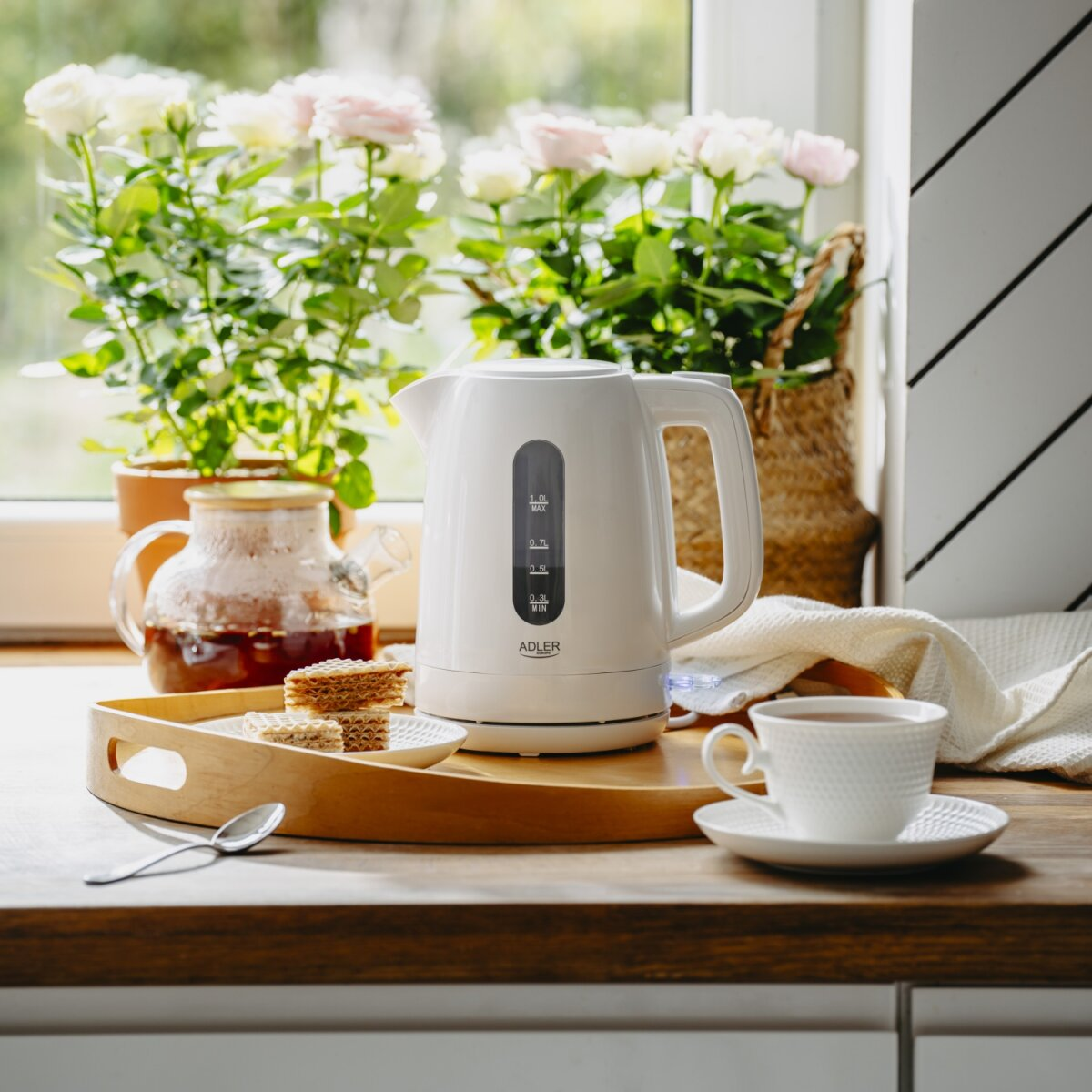 Ein weißer Wasserkocher steht auf einem Holztablett mit Waffeln und einer Teetasse. Pflanzen sind dahinter.