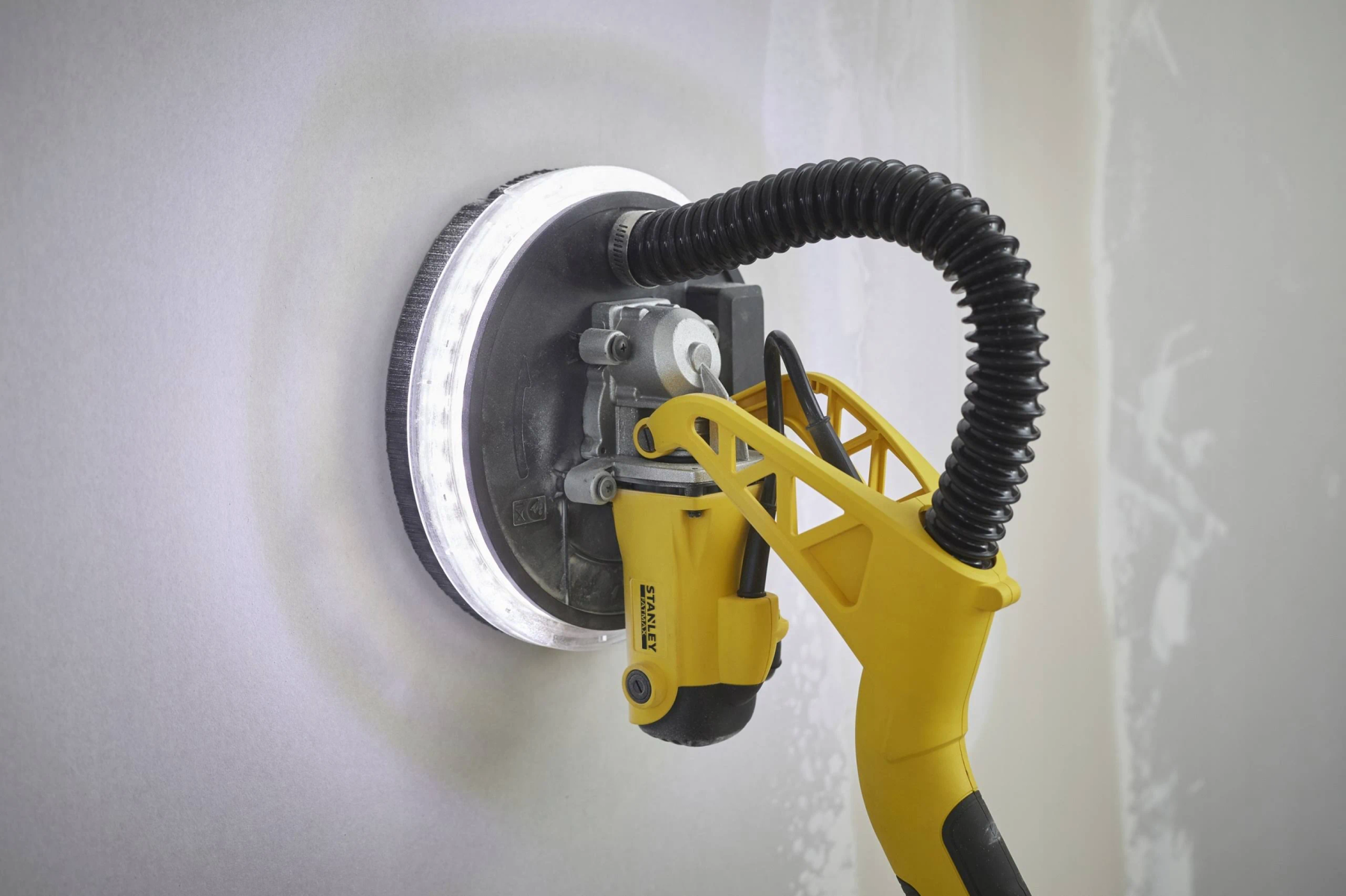 Żółta szlifierka do płyt gipsowo-kartonowych z czarnym wężem szlifuje białą ścianę.