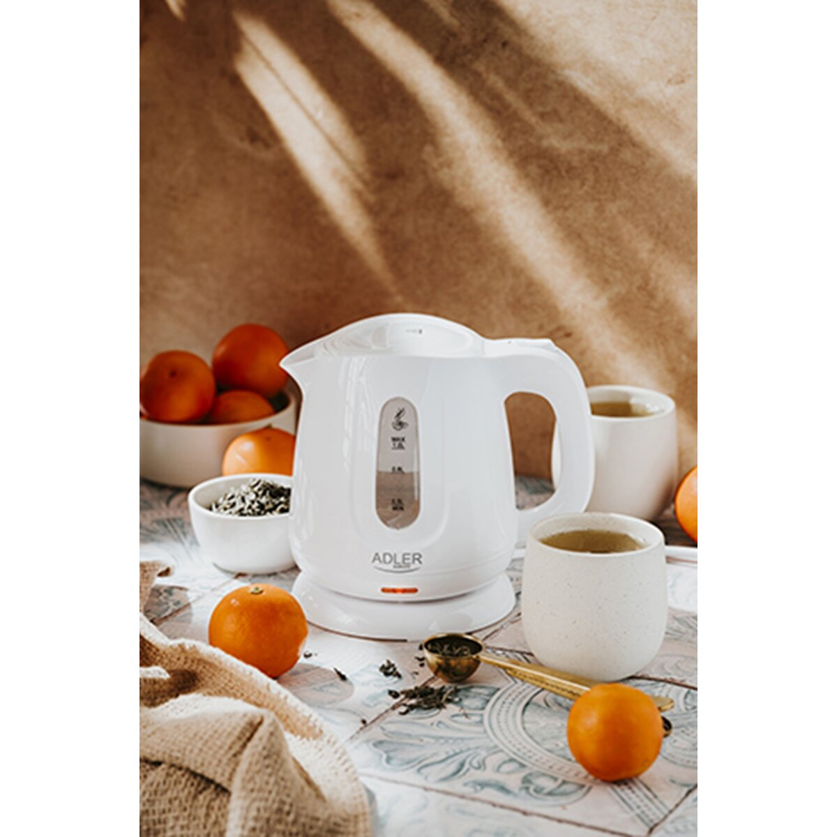 Ein weißer Wasserkocher mit Tassen, Orangen und Tee auf einem gemusterten Tisch.