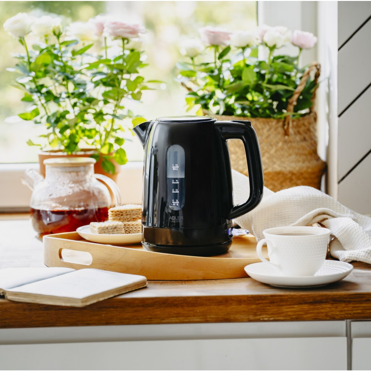 Schwarzer Wasserkocher mit Teekanne, Keksen, Tasse und Buch auf einem Holztisch.
