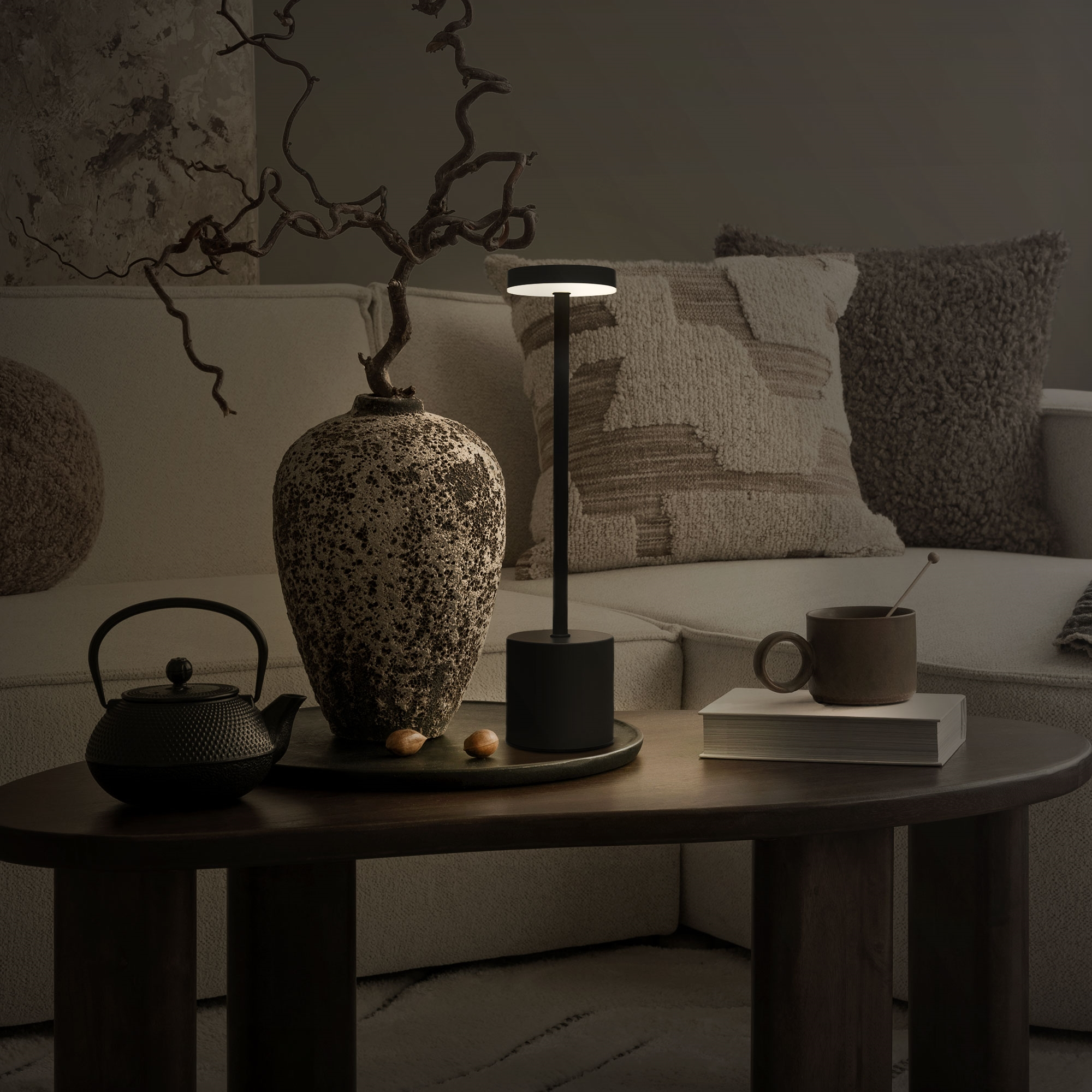 Ein Tisch mit Teekanne, Vase, Lampe und Buch. Auf einem Sofa liegen Kissen.