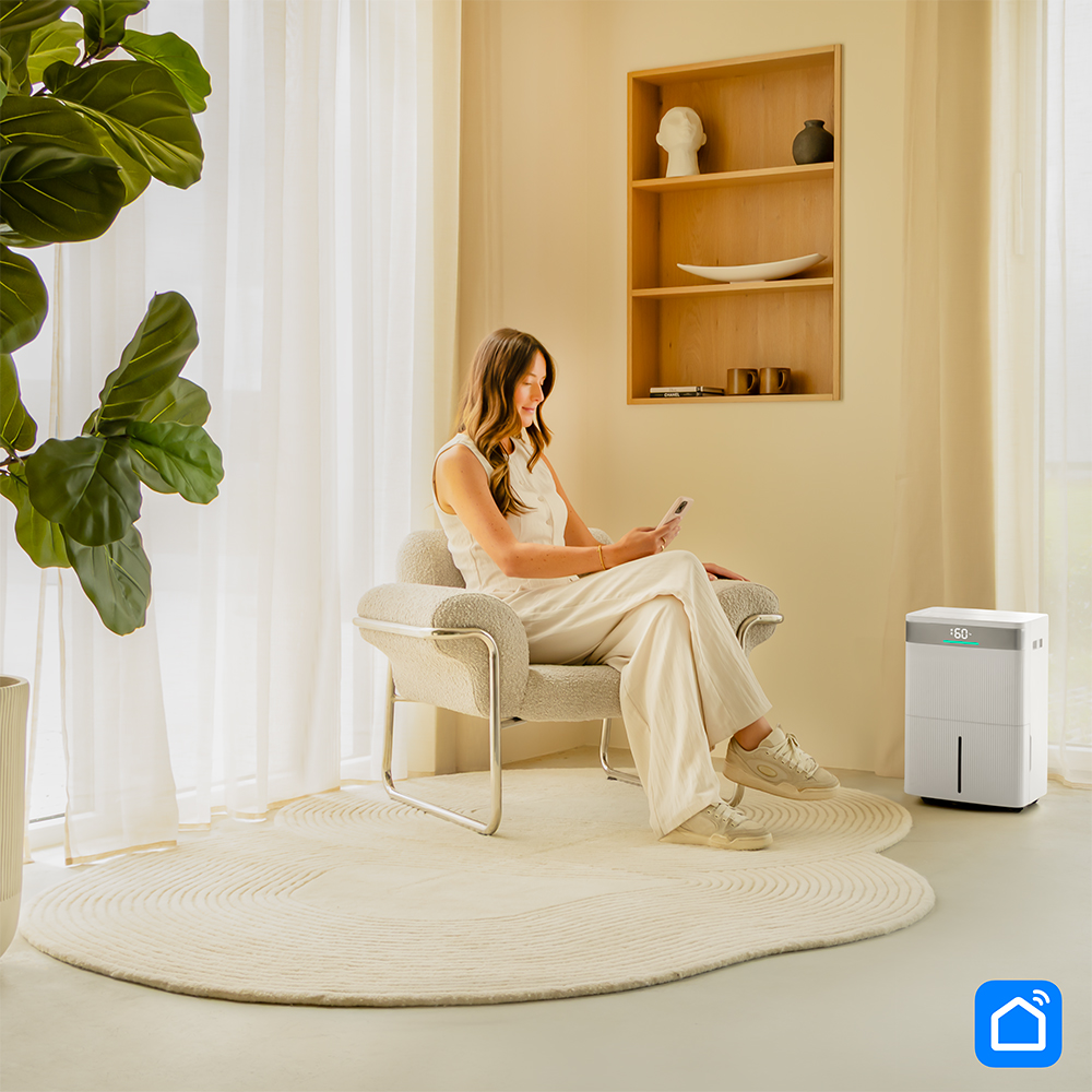 Woman sits in armchair, looking at phone. Dehumidifier is nearby.