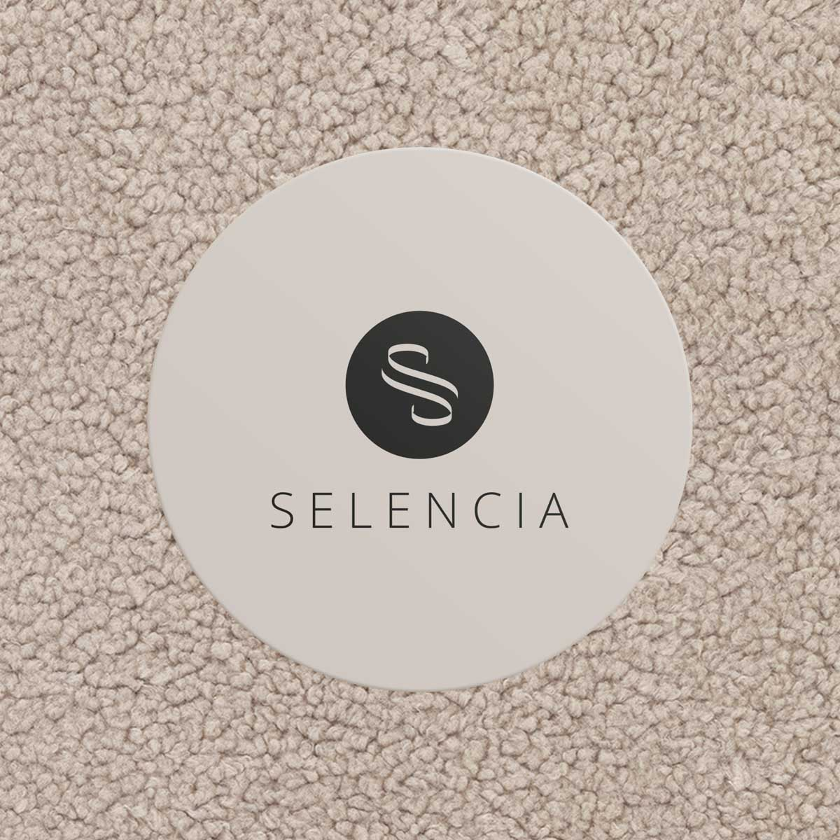 A round coaster with the Selencia logo sits on a light beige carpet.