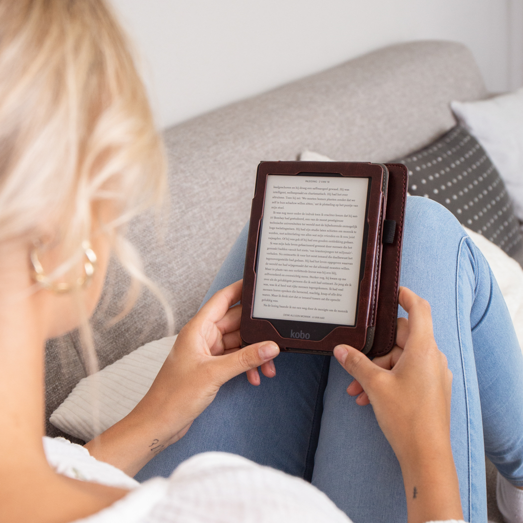 A woman sits on a couch, reading an e-reader with a brown cover.