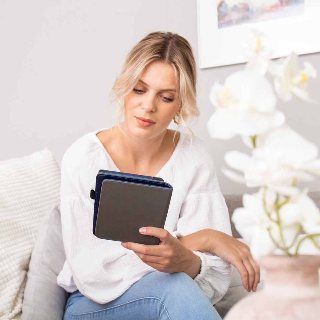 Een blonde vrouw in een witte top zit op een bank en houdt een tablet vast.