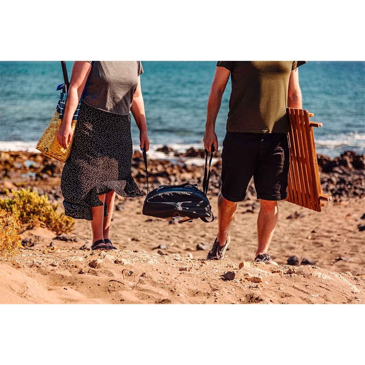 Due persone camminano su una spiaggia, una porta una borsa e una sedia di legno.