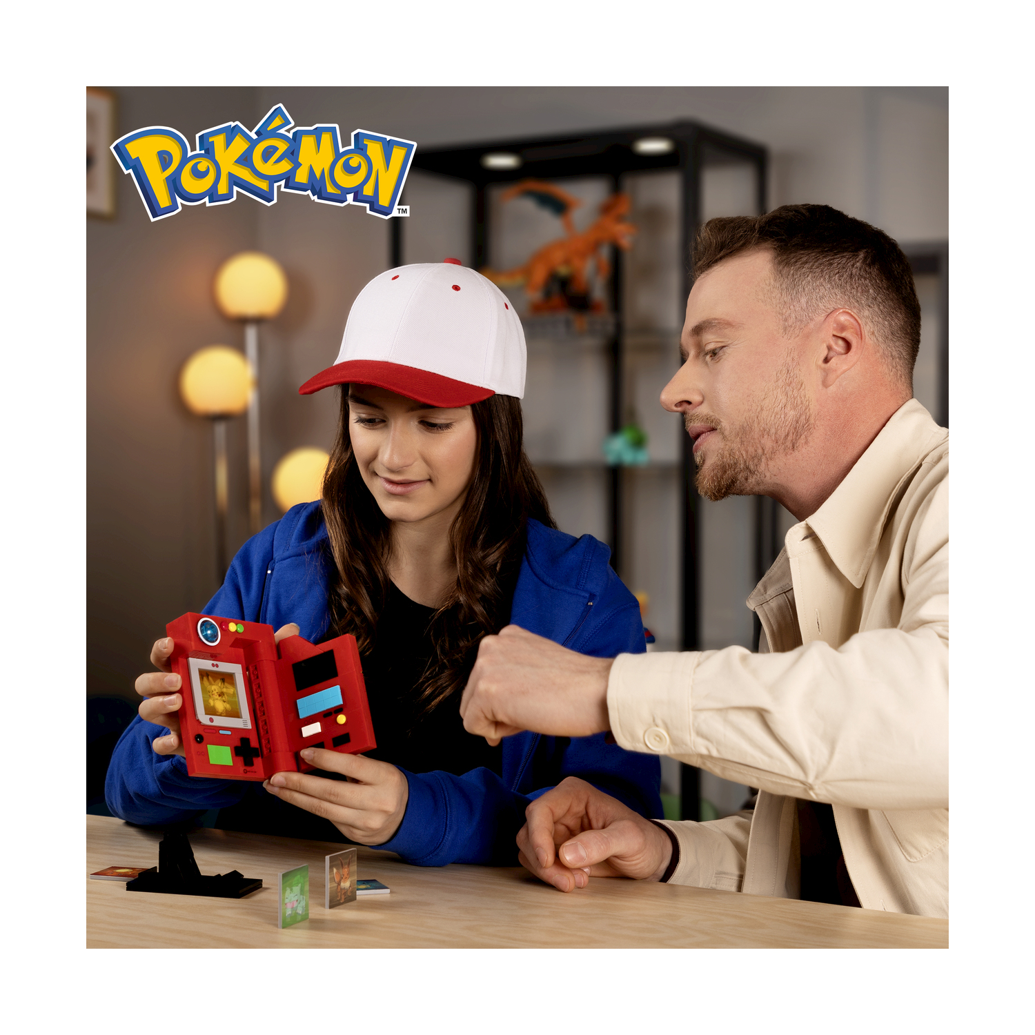 Dwie osoby bawią się czerwonym urządzeniem Pokémon i kartami kolekcjonerskimi przy drewnianym stole.