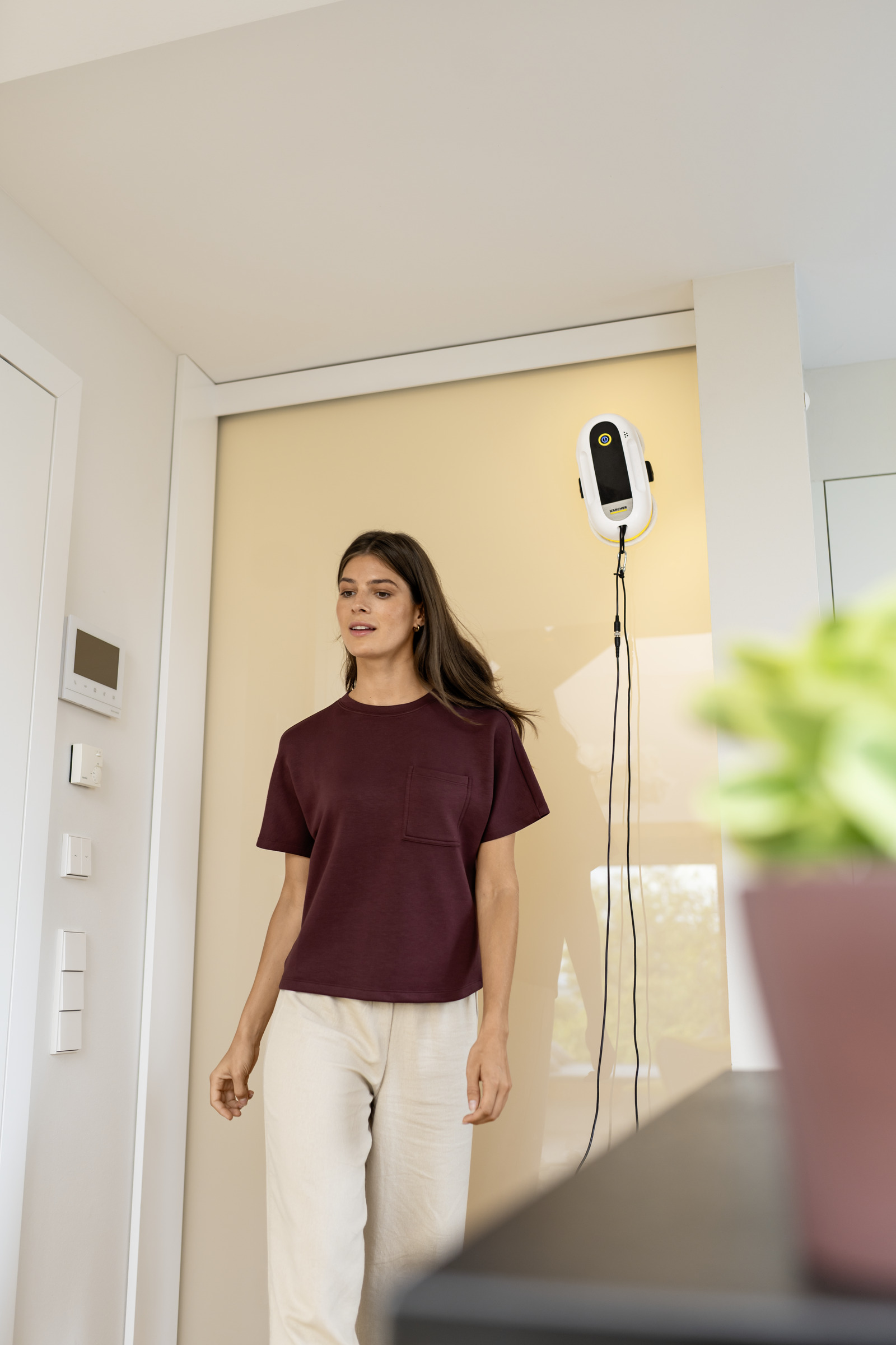 Frau in bordeauxfarbenem Oberteil und beiger Hose neben Fensterreinigungsroboter.