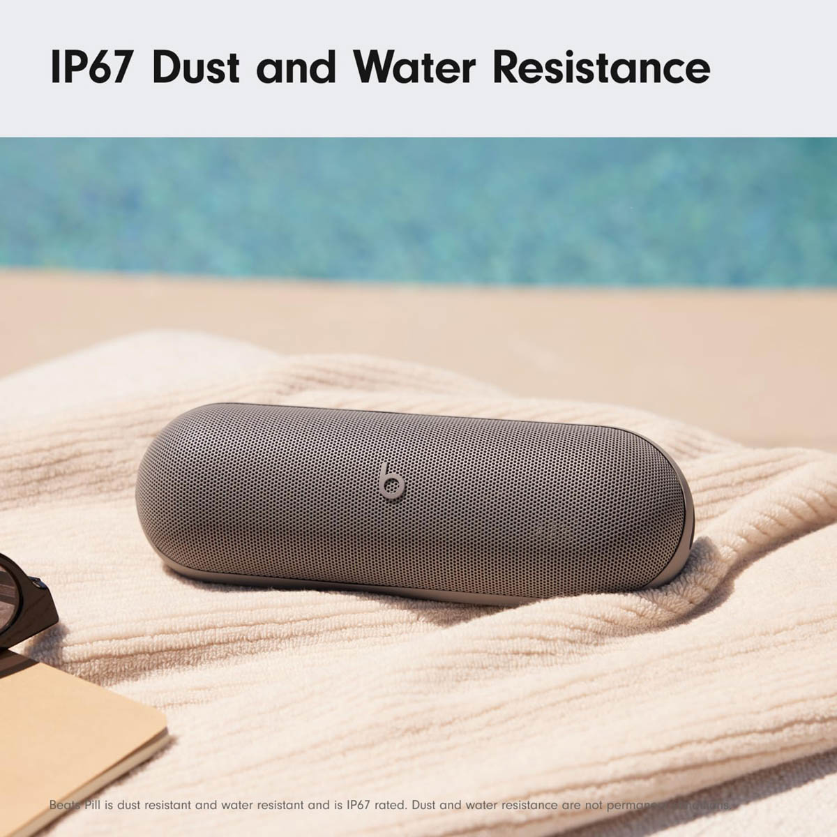 Grauer Beats Pill Lautsprecher liegt auf einem Handtuch, Pool im Hintergrund.