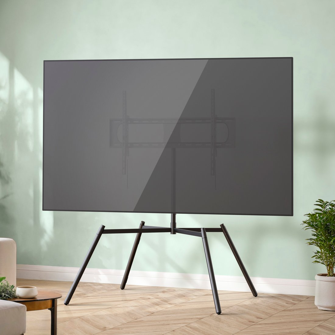 Duży, ciemny telewizor stoi na czarnym stojaku przed jasnozieloną ścianą.