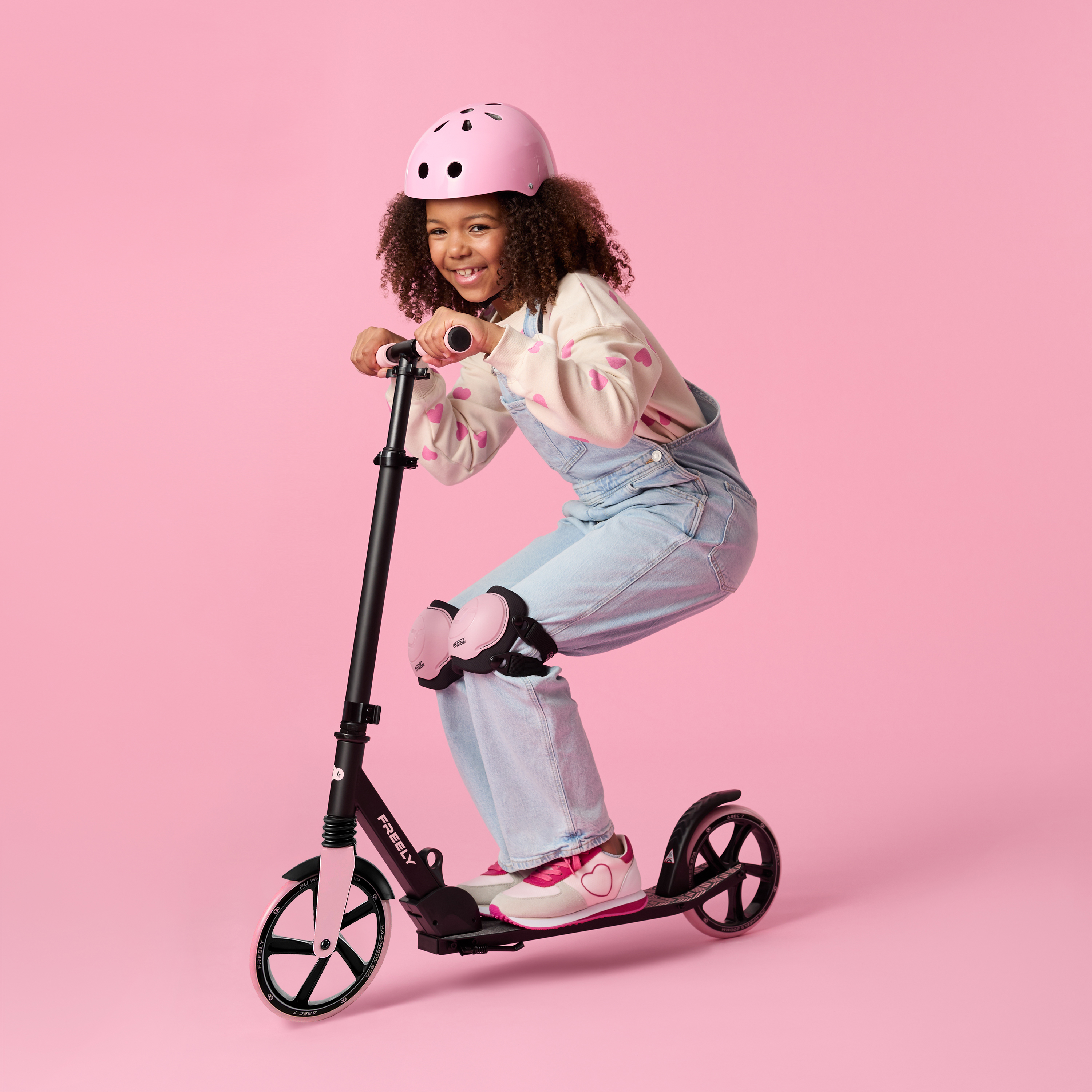 Ein Mädchen mit rosa Helm fährt Roller, trägt Latzhose und Schutzpolster.