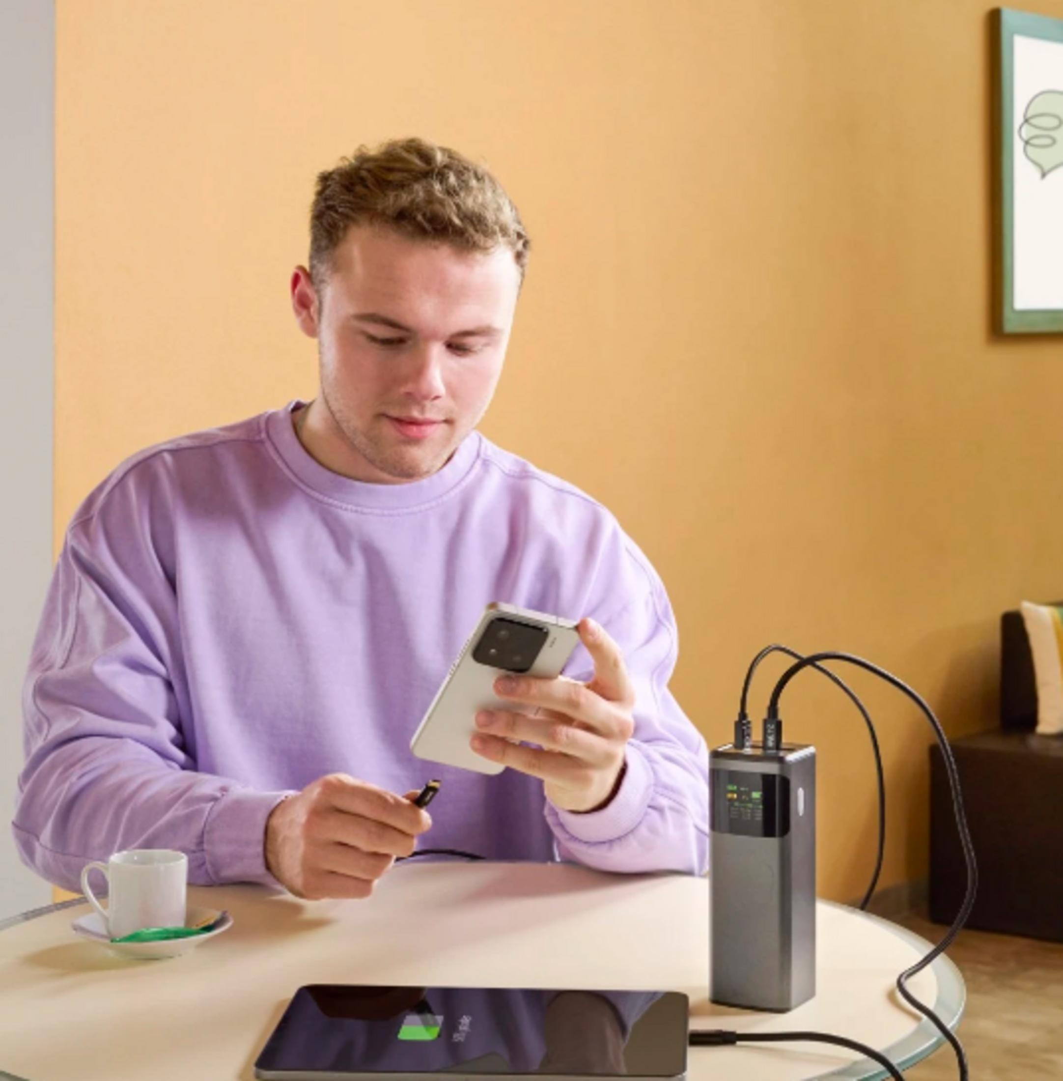 Mann lädt Handy mit Powerbank, Tablet und Kaffeetasse auf dem Tisch.