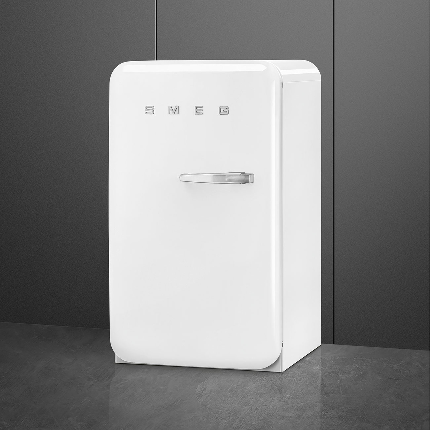 Un refrigerador SMEG blanco con tirador cromado sobre suelo oscuro y pared oscura.