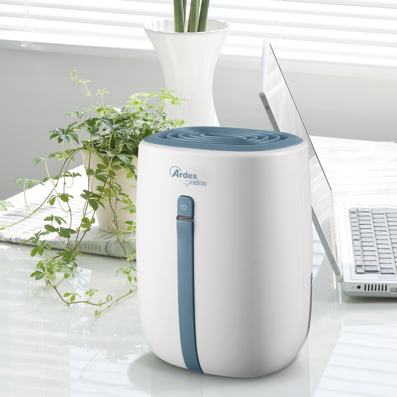 Biały oczyszczacz powietrza stoi na biurku obok zielonej rośliny i laptopa.