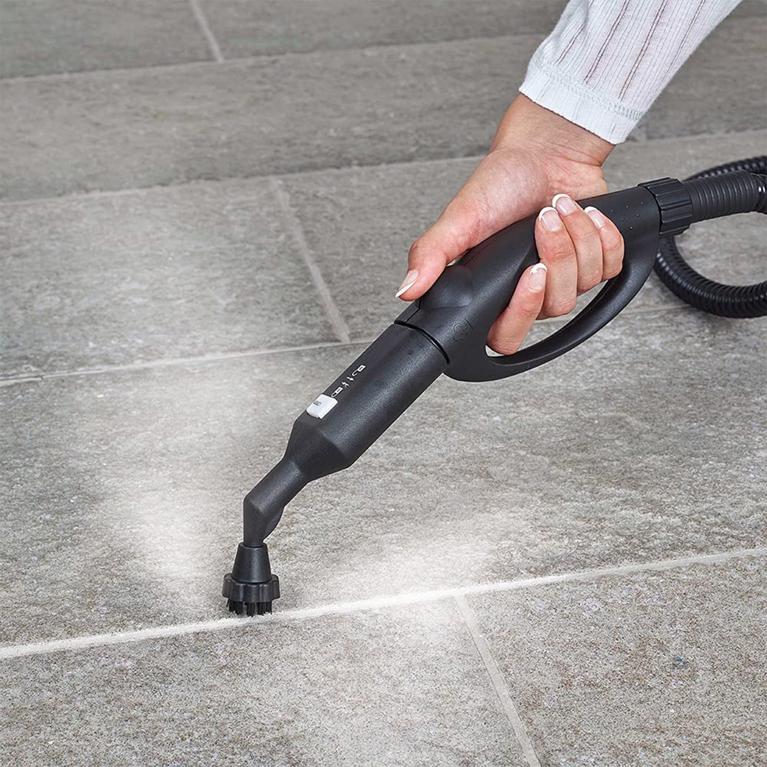 Ręka trzyma czarny odkurzacz parowy rozpylający parę na szare płytki.