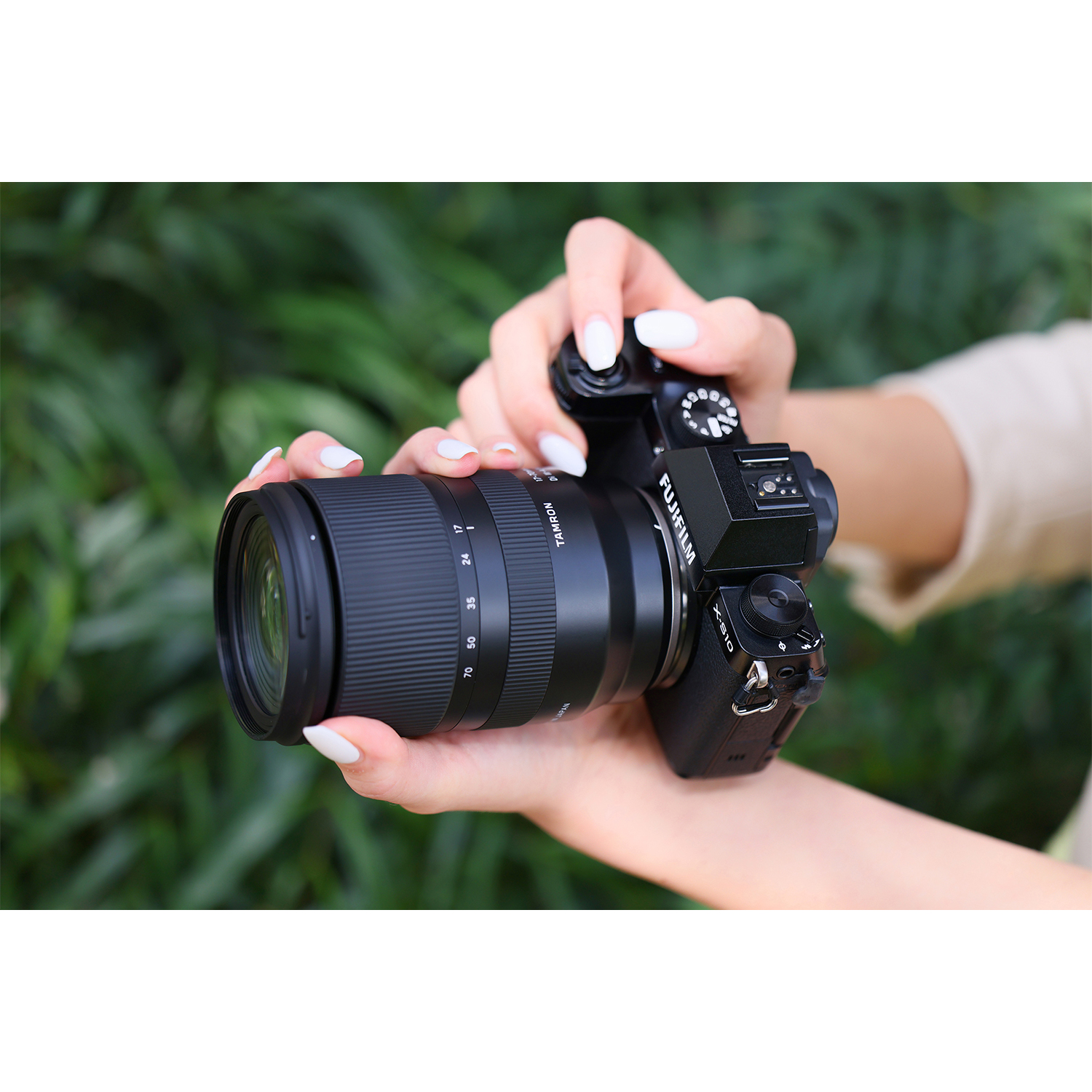 Dłonie trzymają czarny aparat z obiektywem, tło zielonych liści.