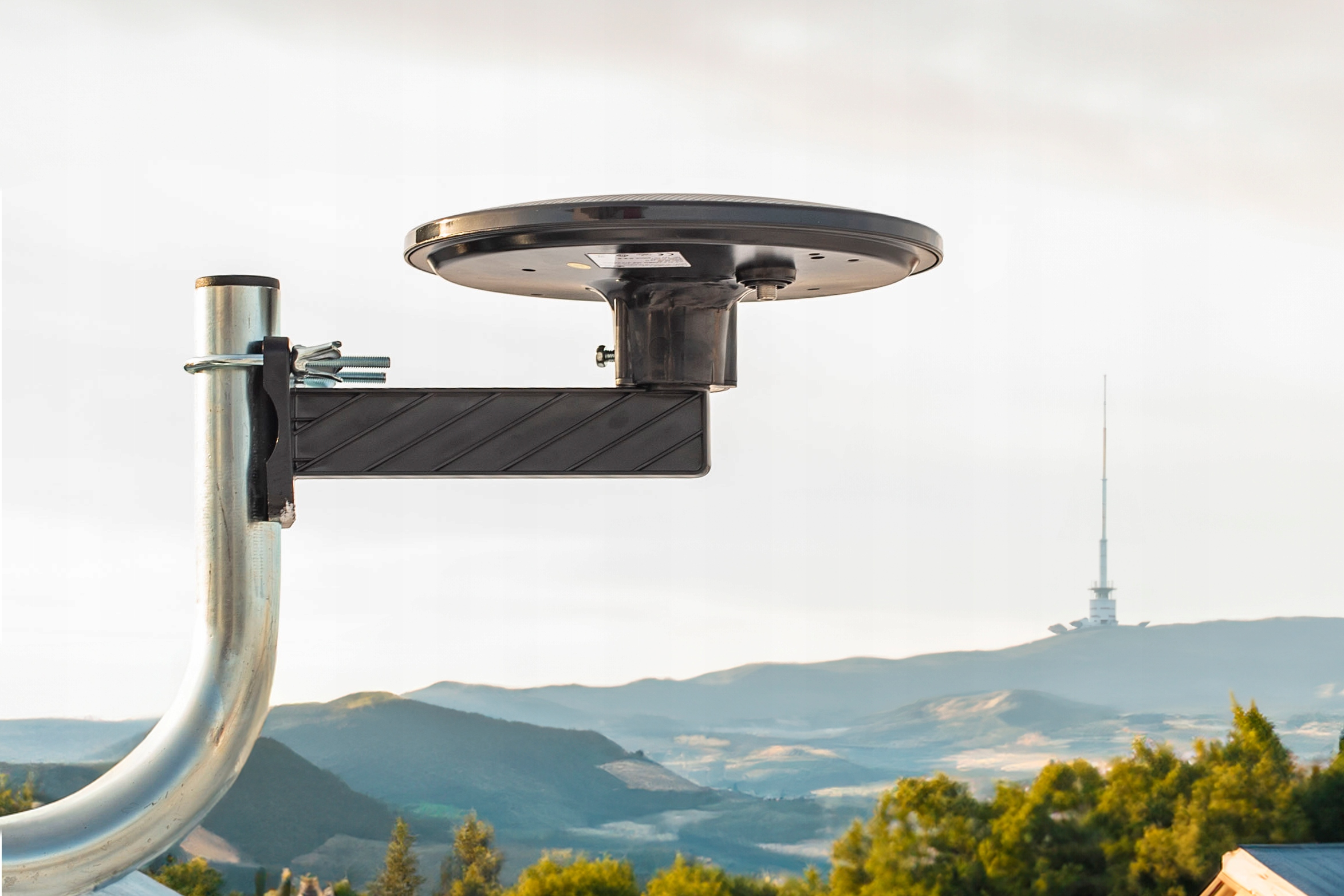 Uchwyt anteny satelitarnej na tle pagórkowatego krajobrazu i wieży.
