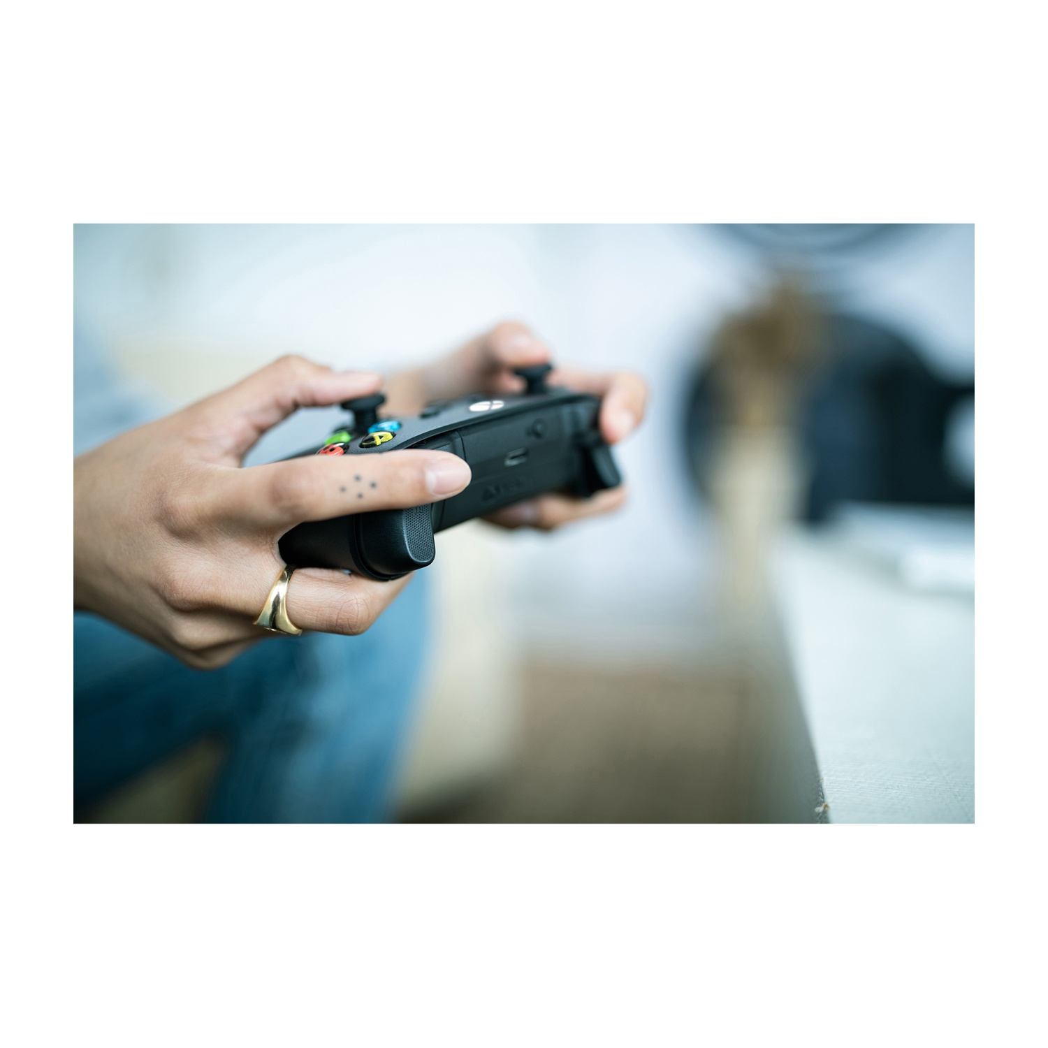 Dłonie trzymające czarny kontroler, kciuki na przyciskach. Złoty pierścionek na palcu.