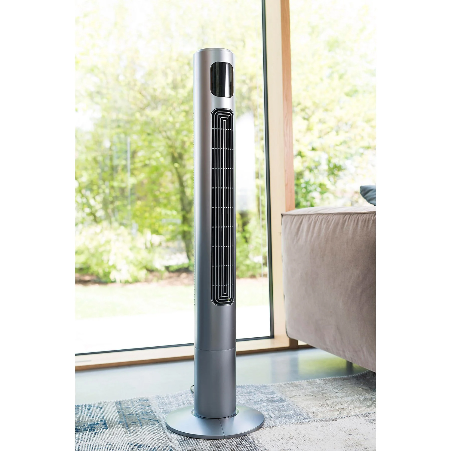 Ein schlanker, silberner Turmventilator steht auf einem Teppich vor einem großen Fenster.