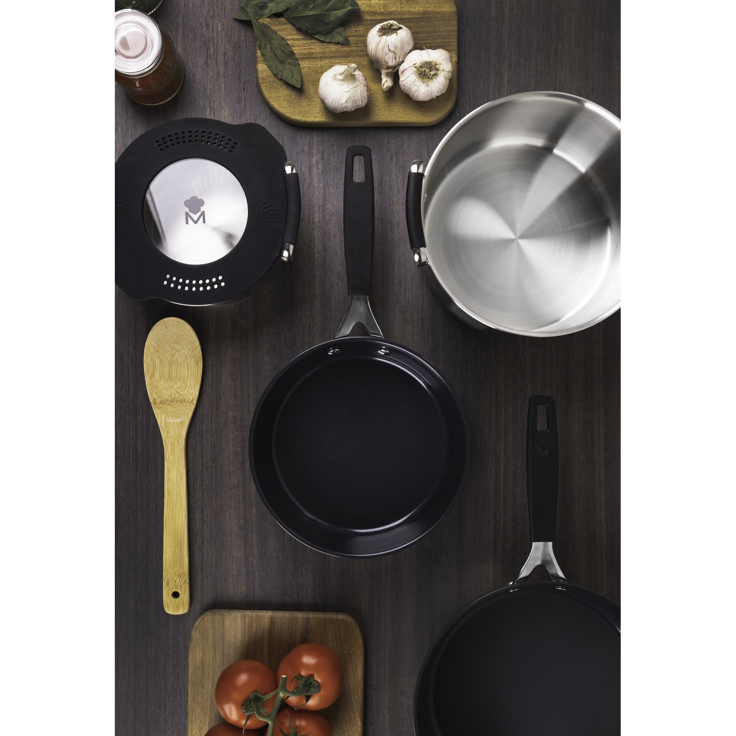 Utensilios de cocina con ajo, tomates y hierbas en mesa de madera.