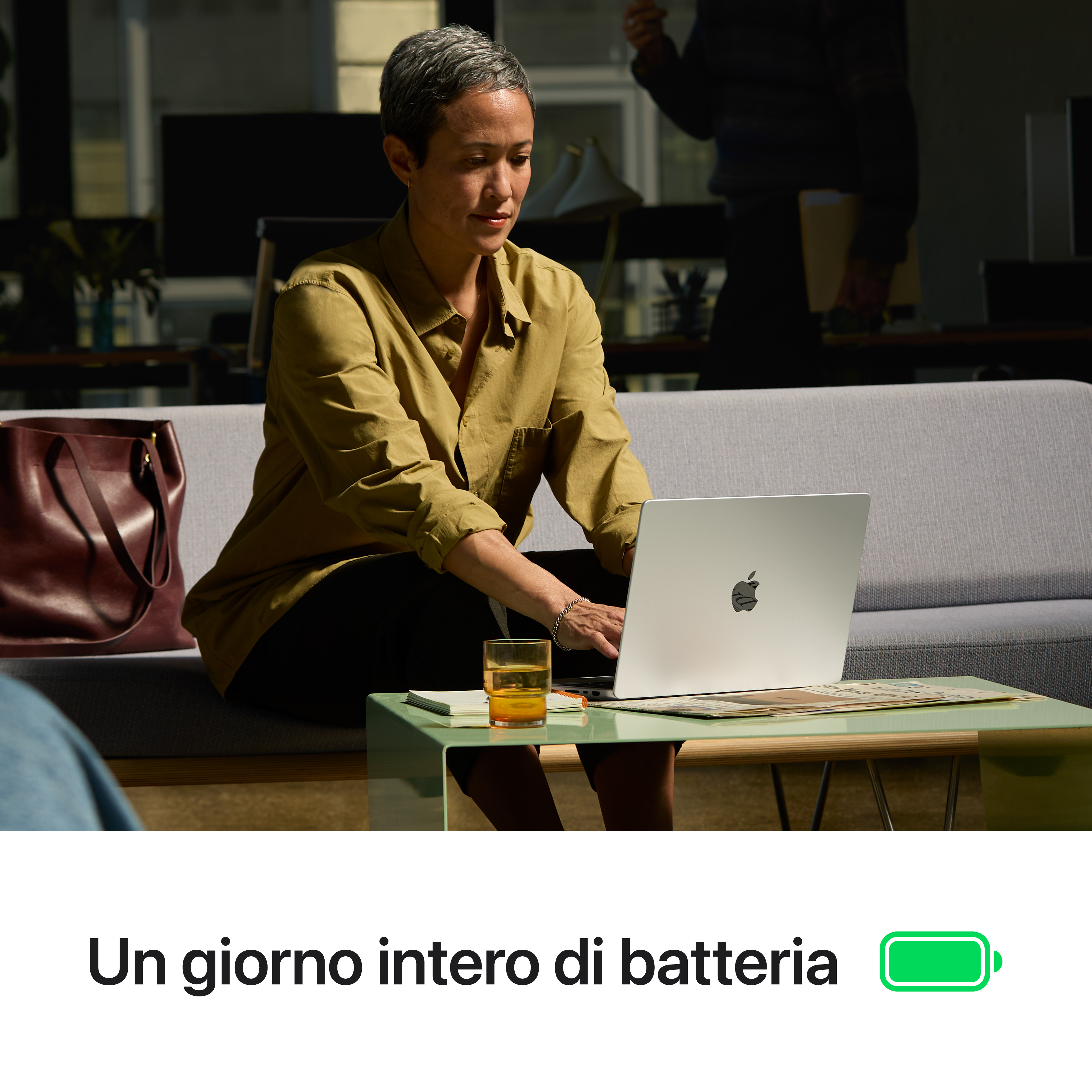 Donna lavora al laptop, vicino bicchiere d'acqua e giornali.