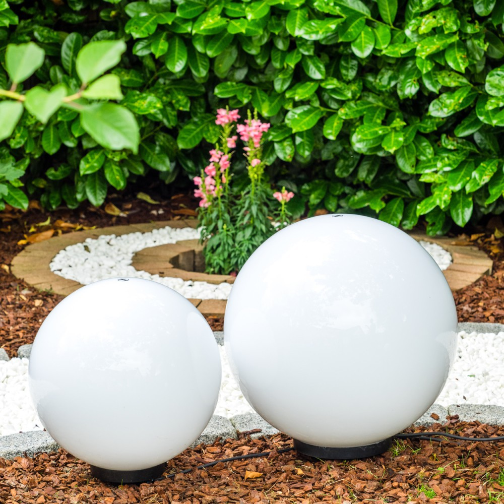 Zwei weiße Kugelleuchten in einem Garten mit grünen Büschen und rosa Blumen.