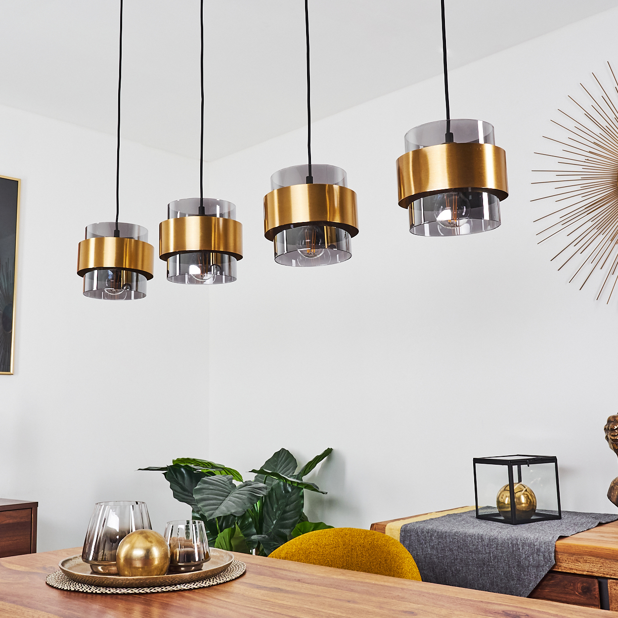Vier moderne Hängeleuchten mit goldenen und rauchfarbenen Zylindern hängen über einem Holztisch.