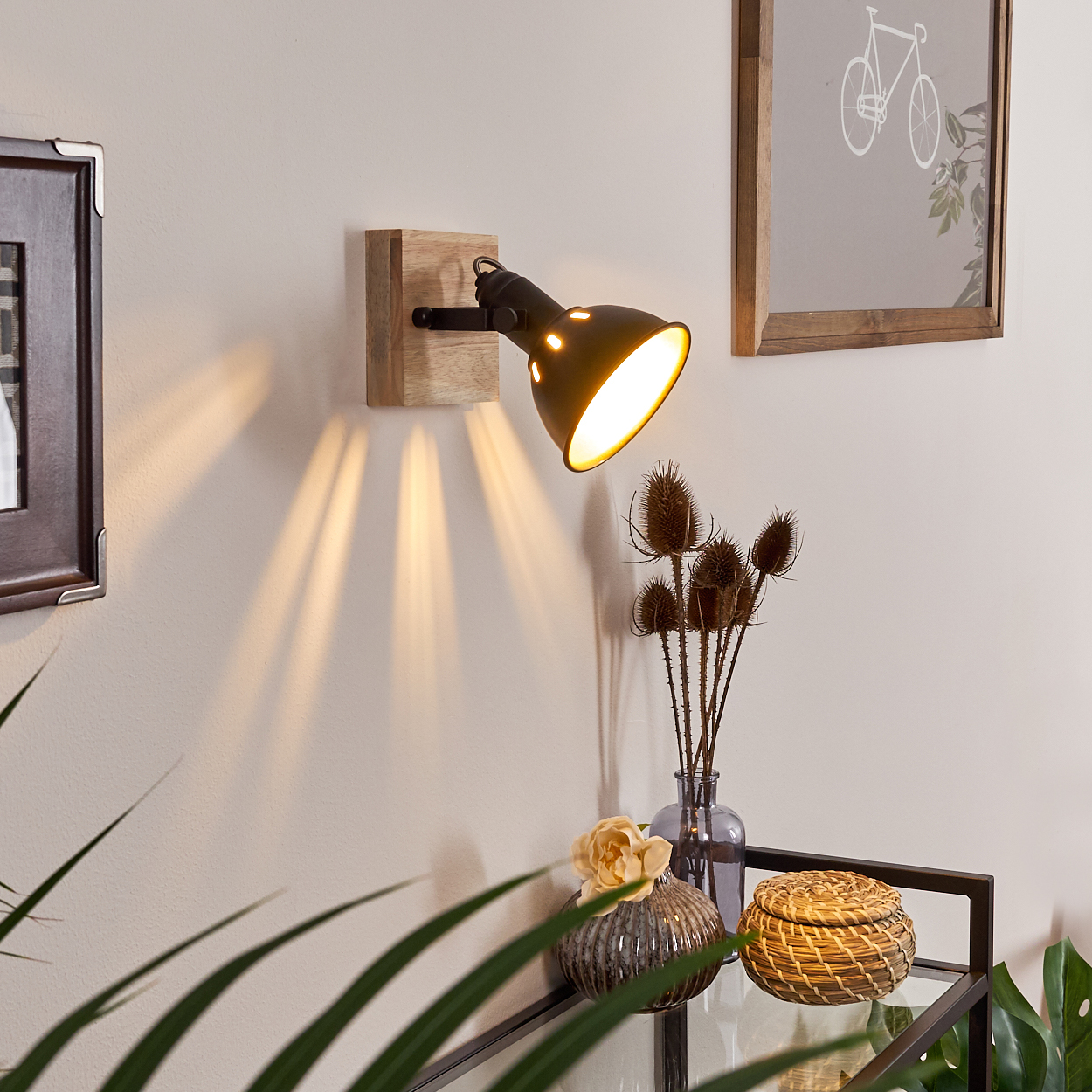 Ein schwarzer Wandstrahler beleuchtet eine Wand, daneben ein Bild und Trockenblumen.