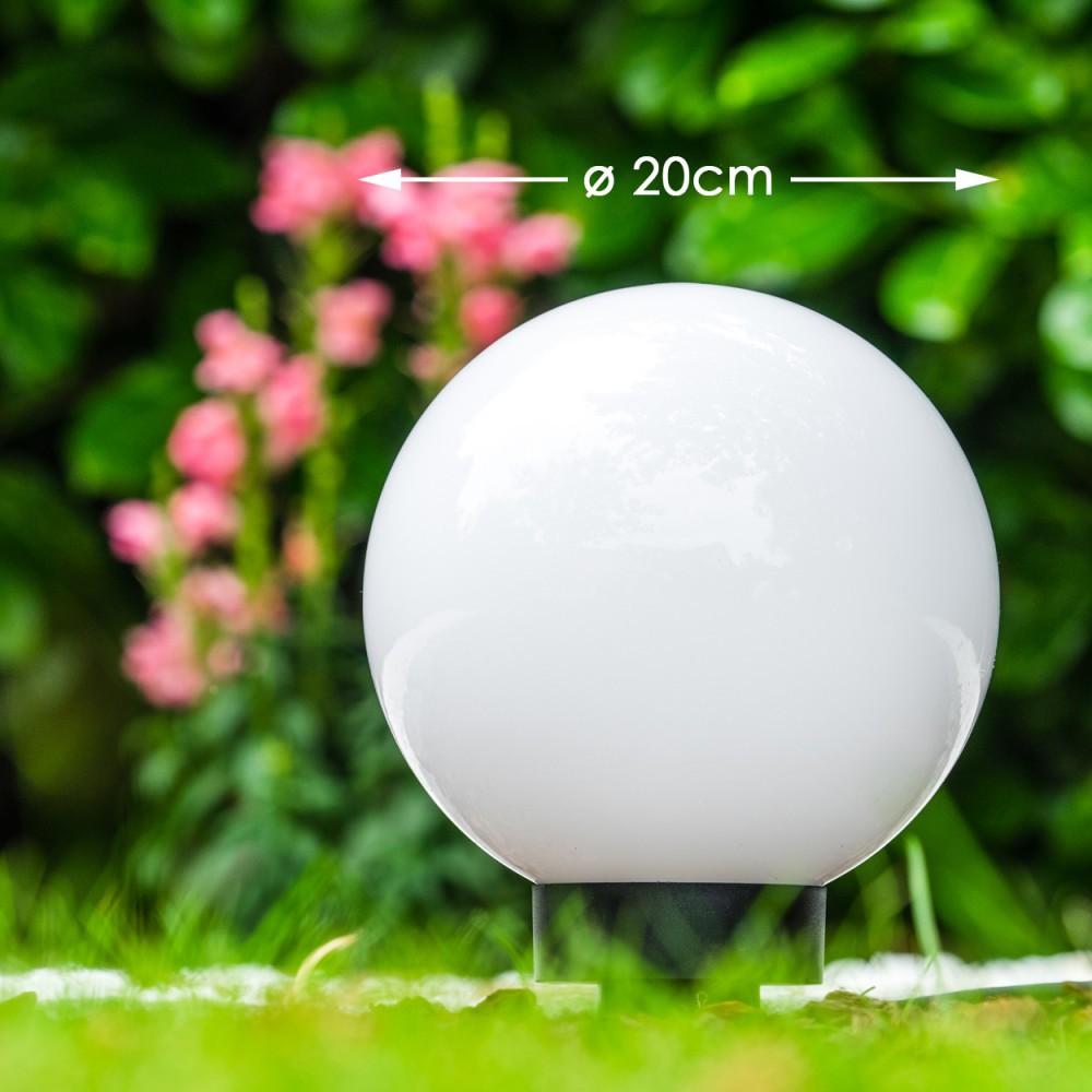 Eine weiße Glaskugel Lampe auf einem Sockel im Gras mit rosa Blumen dahinter.