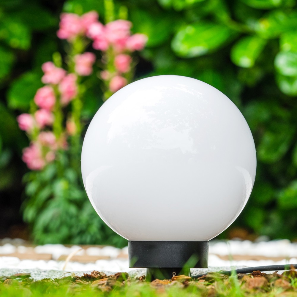 Eine weiße kugelförmige Lampe steht im Gras vor rosa Blumen und grünem Laub.